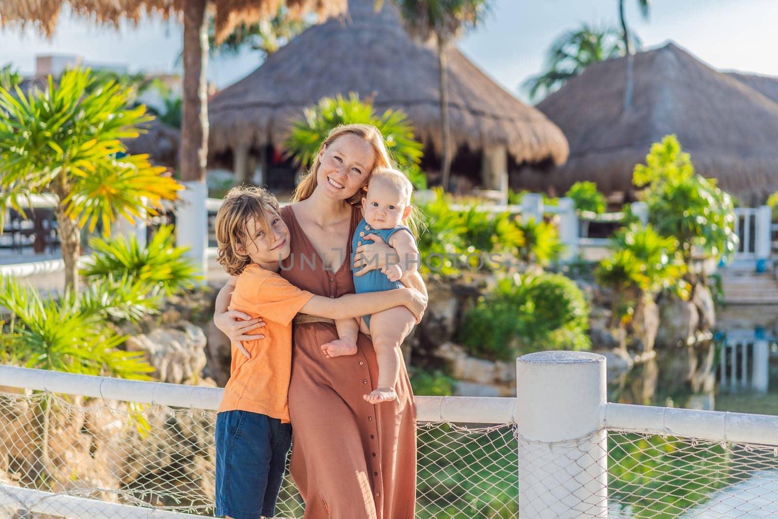 In a tropical paradise, a mom lovingly embraces her baby and 10-year-old son amid lush palm trees and thatched roofs, creating heartwarming family memories.