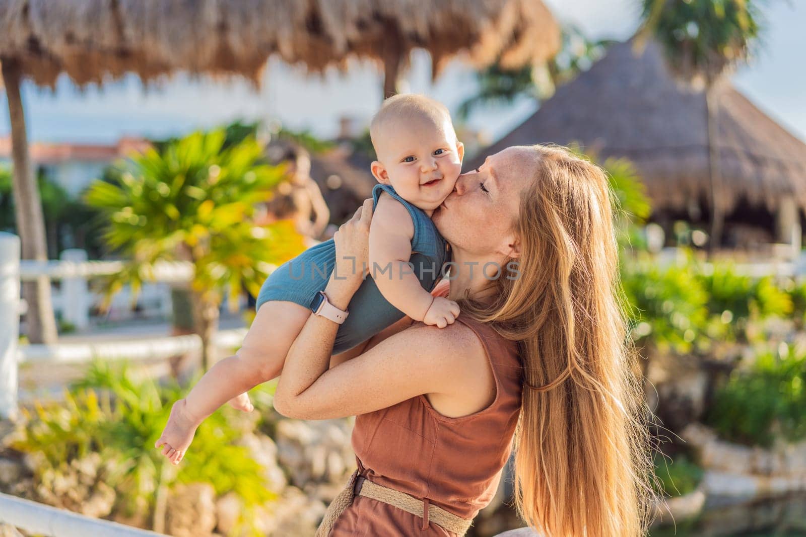 Amidst tropical palms and thatched roofs, a loving mom embraces her baby, sharing warmth and affection in a tranquil outdoor setting by galitskaya