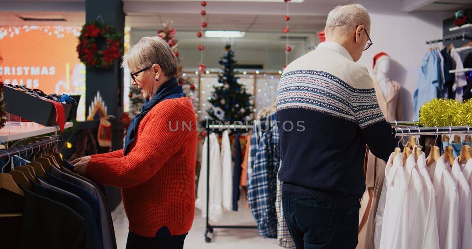 Octogenarian clients browsing through clothing racks in festive ornate fashion shop during winter holiday season. Senior couple happy after finding colorful blazers to gift at Xmas family gathering