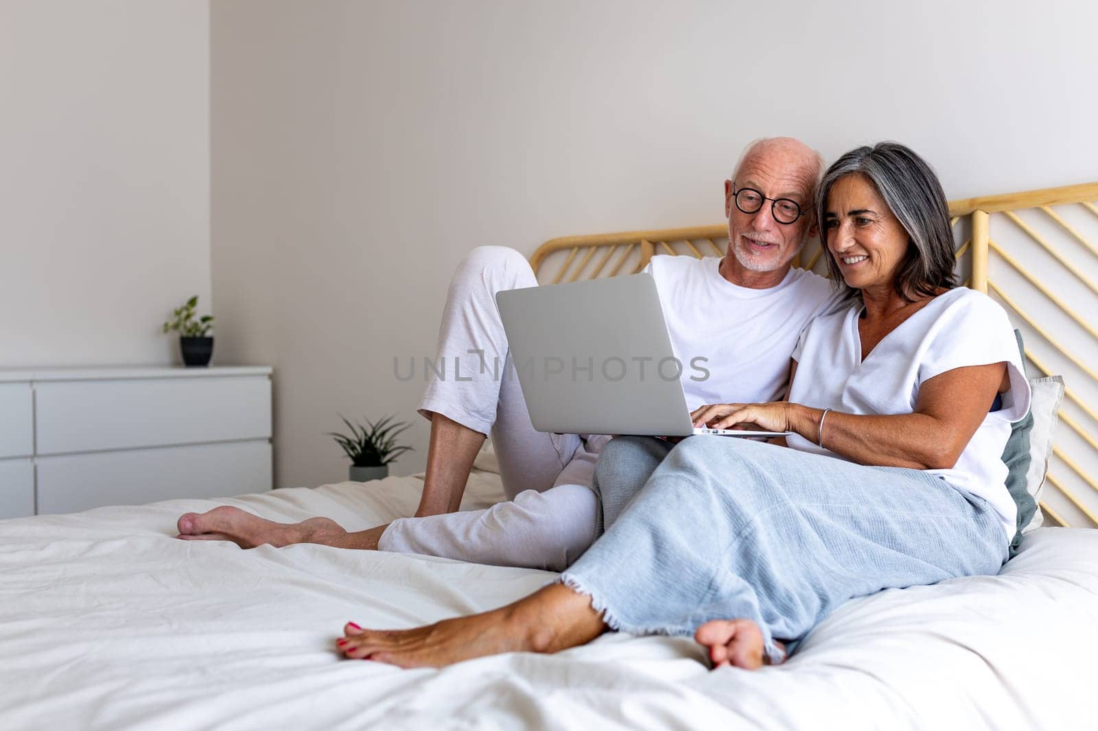 Happy mature couple using laptop together relaxing on bed. Copy space. Lifestyle and technology concepts.