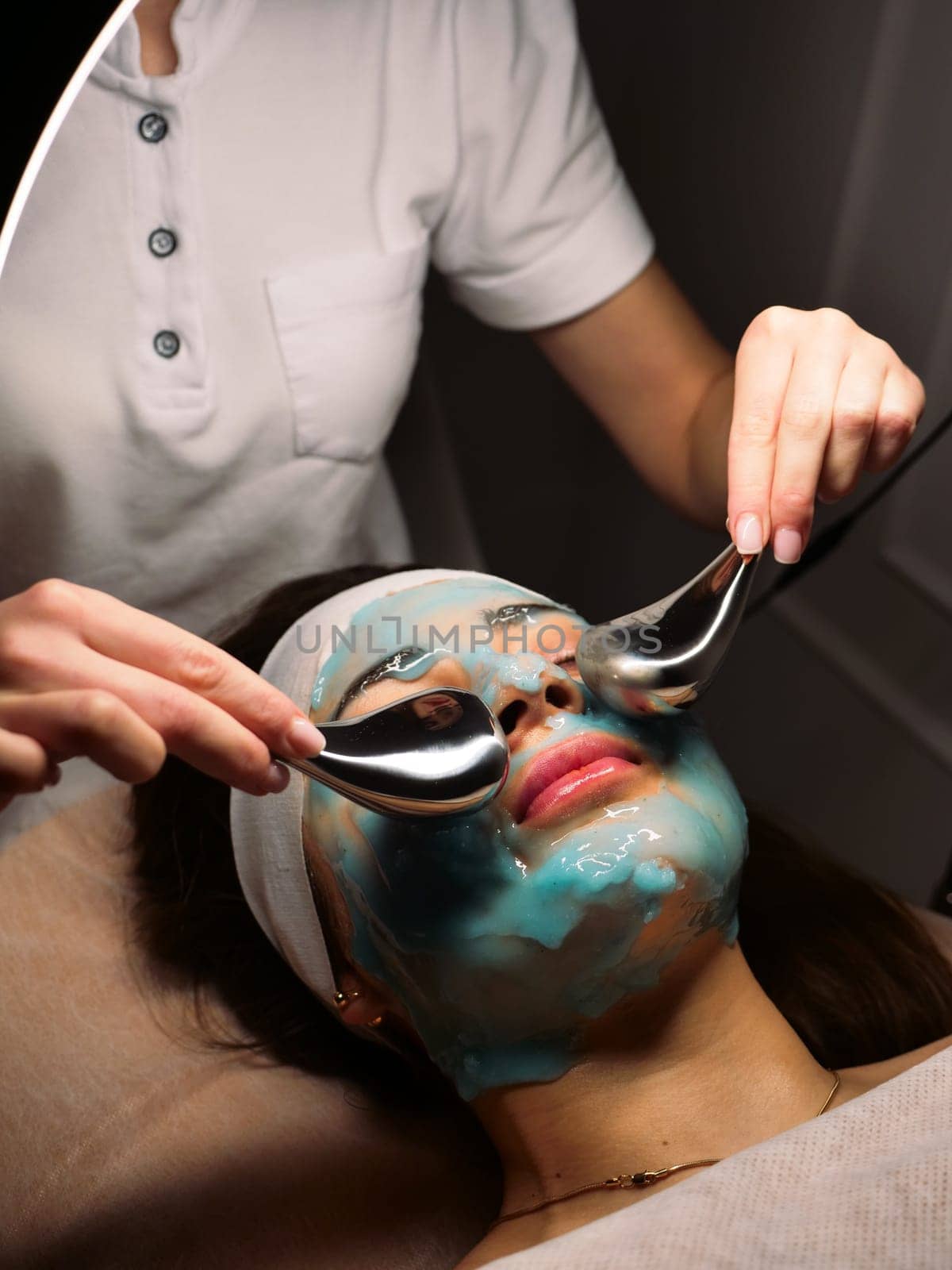 Cosmetologist does cryo massage with globules on a blue hydrogel alginate mask in the beauty salon, vertically, close-up. Woman Facial Skin & Body Care
