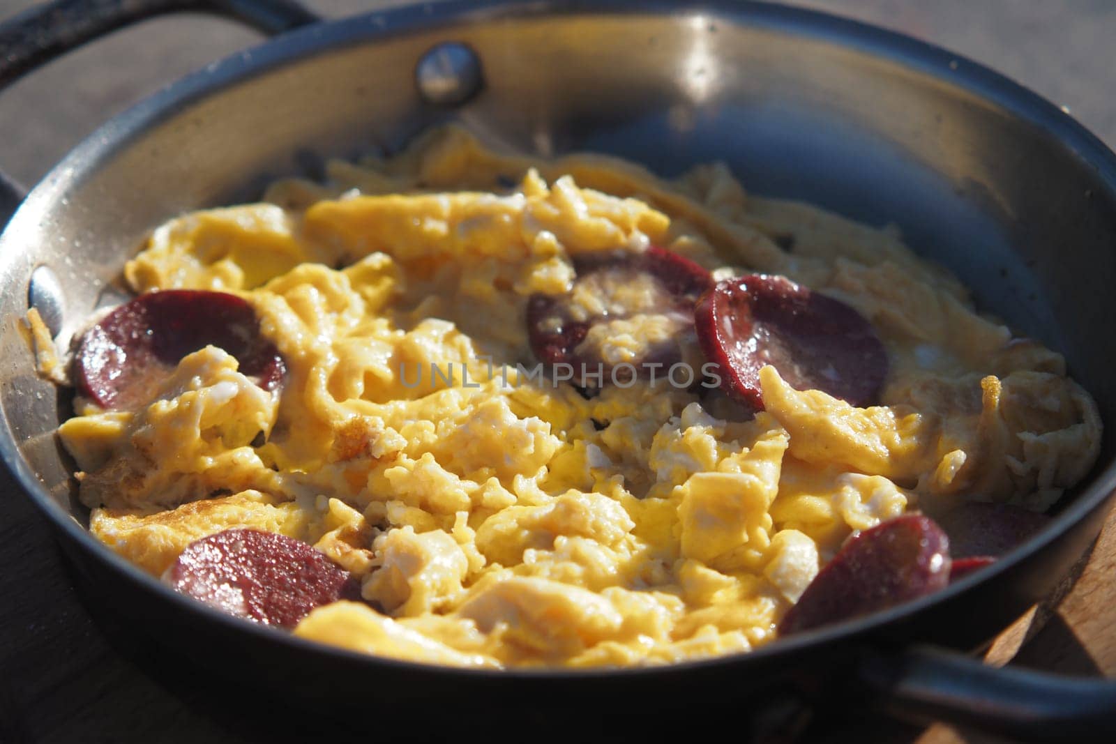 A skillet filled with fluffy scrambled eggs mixed with slices of sausage sits on a wooden surface under clear blue skies. The meal is prepared outdoors, showcasing a relaxed cooking experience.