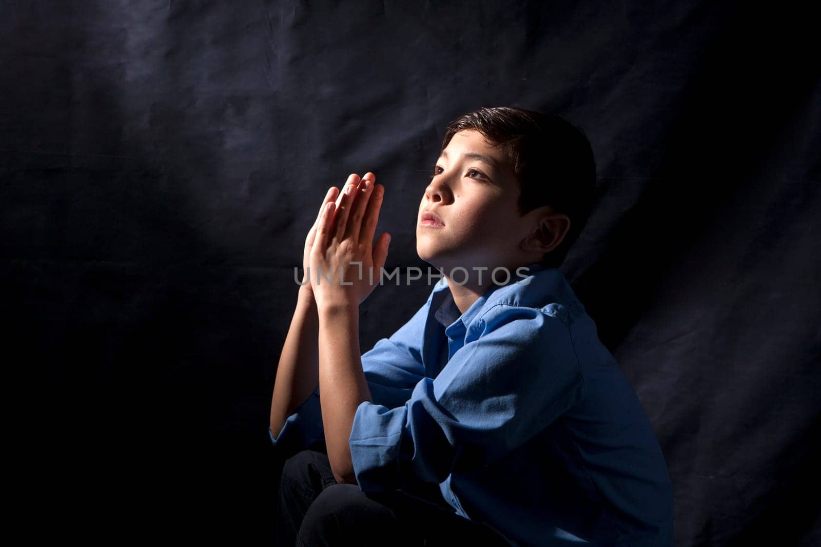 A young boy folds his hands and raises his head in prayer to God.
