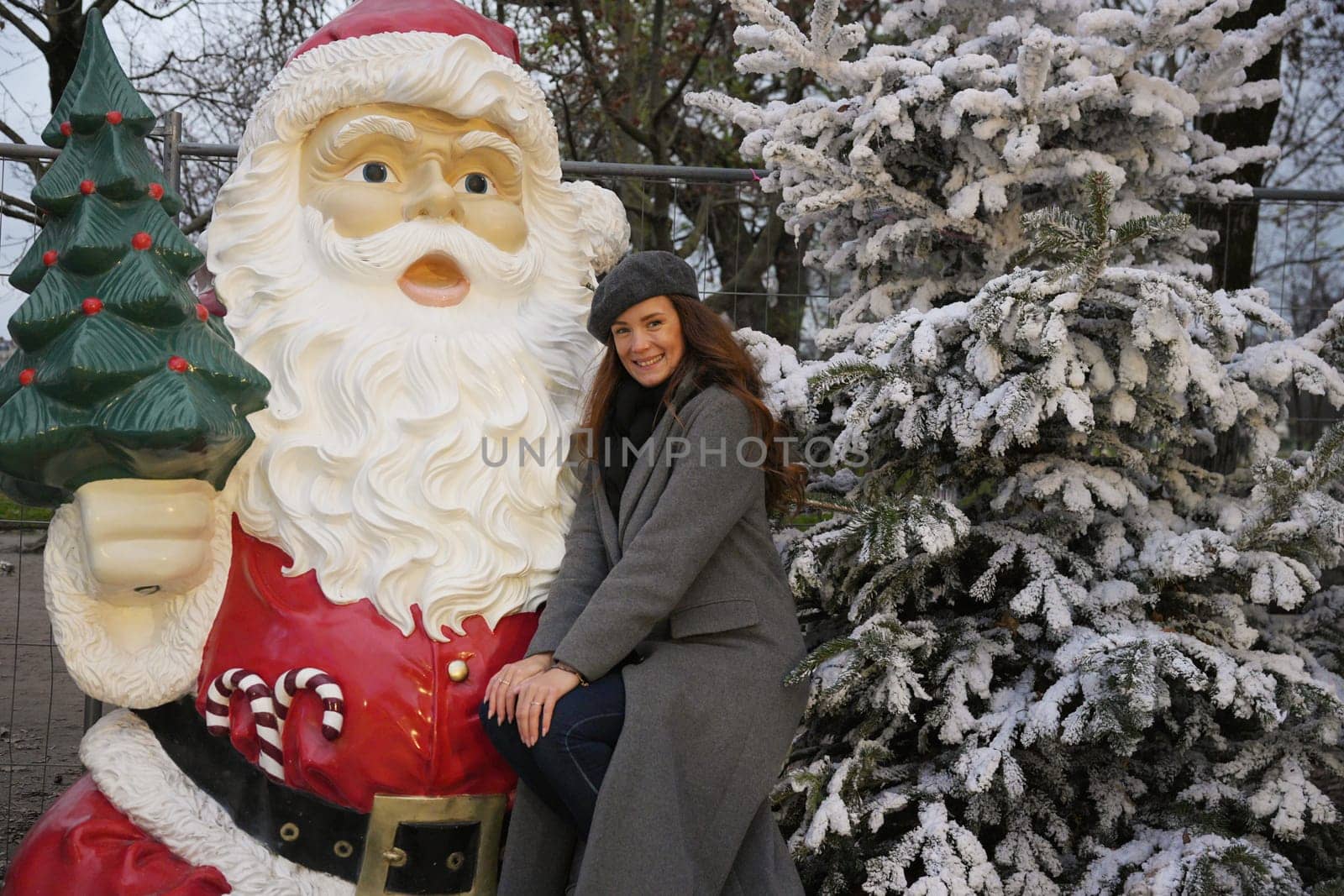 A cheerful woman in a gray coat and beret leans next to a Santa statue and a snow-covered tree, enjoying holiday festivities