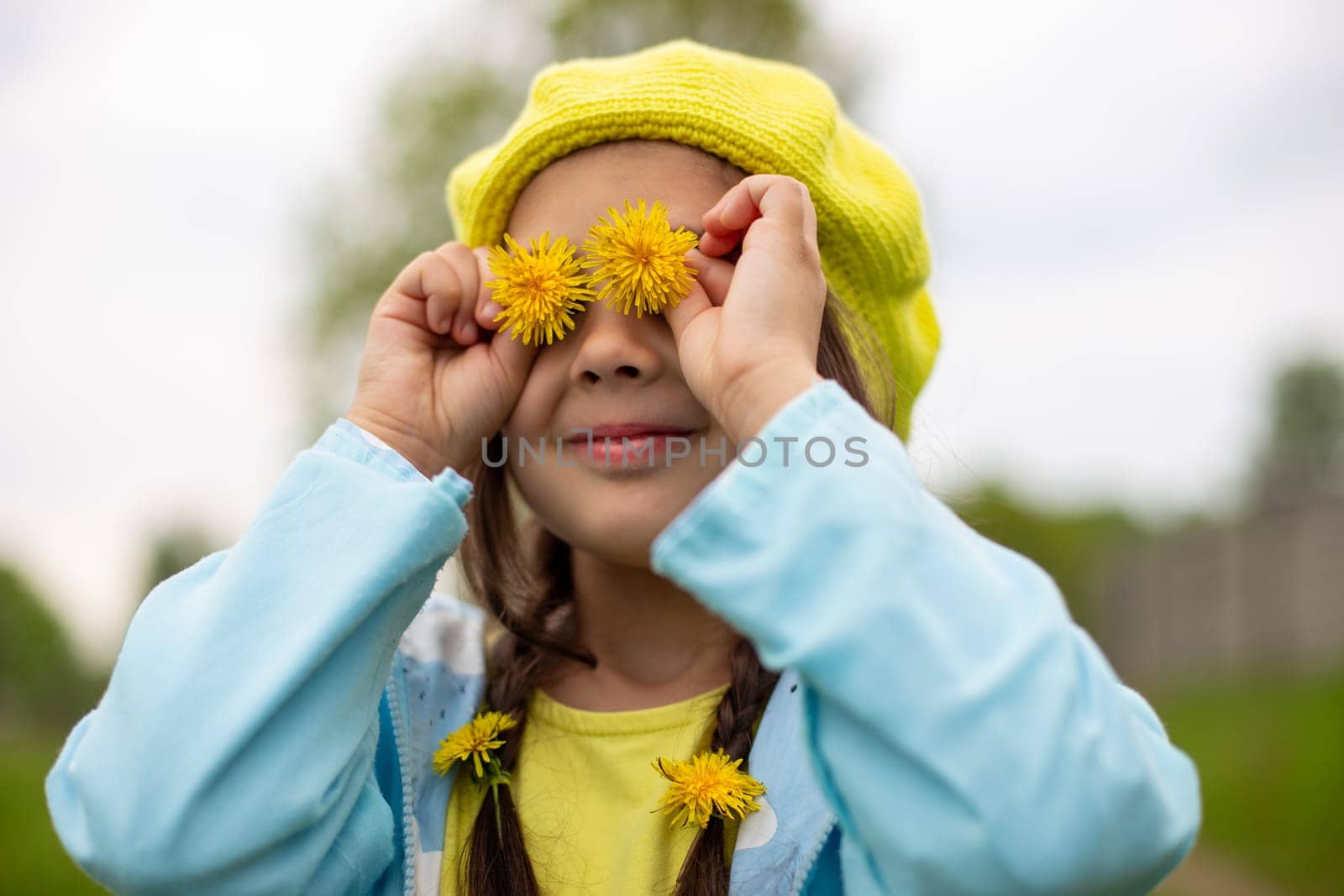 Portrait of a funny charming little girl with two pigtails, in a yellow knitted beret, a blue raincoat, with two yellow dandelion flowers near her eyes stands on nature in spring