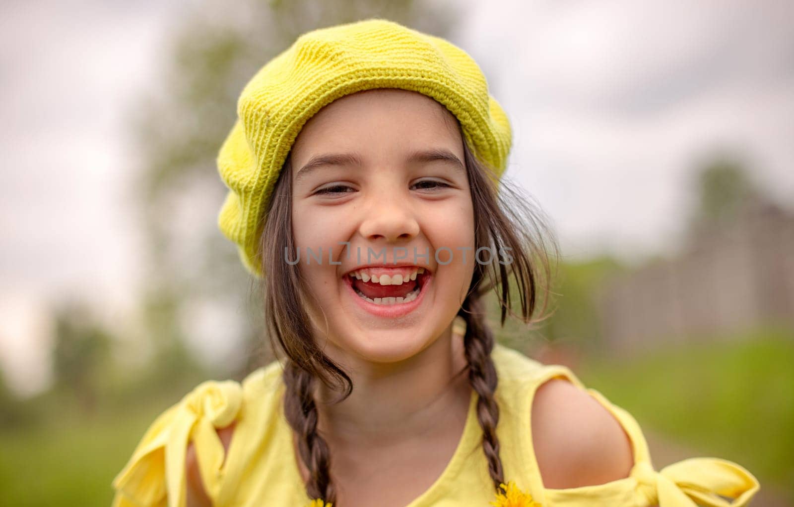 Outdoor shot portrait of a little adorable happy girl in a yellow knitted beret and yellow dress looking straight into the camera and laughing. Happy childhood concept.