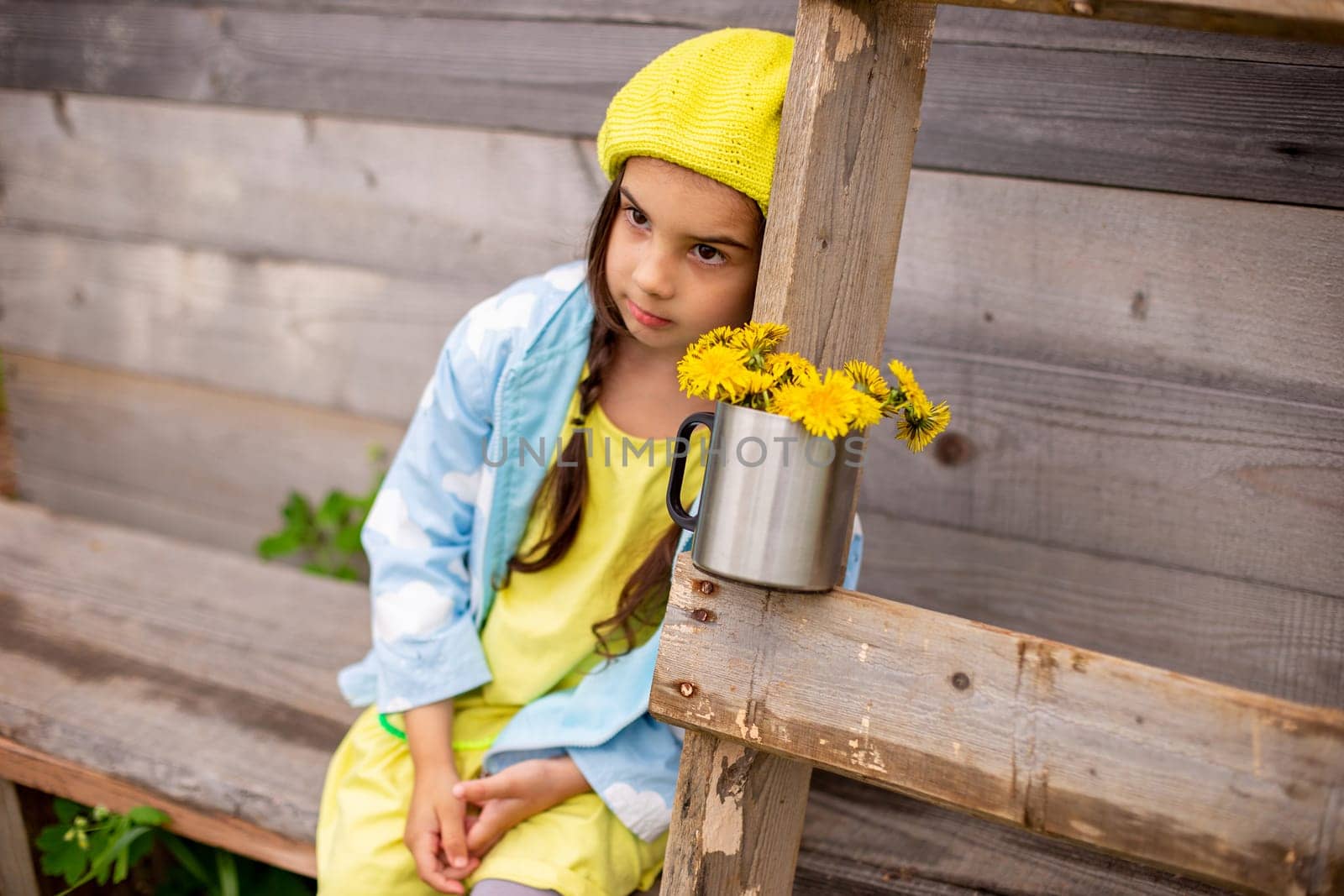 Portrait of a sad adorable little girl with two pigtails, in a yellow beret, blue raincoat, sits in the countryside near a old wooden building with a ladder and a bouquet of dandelions in a mug