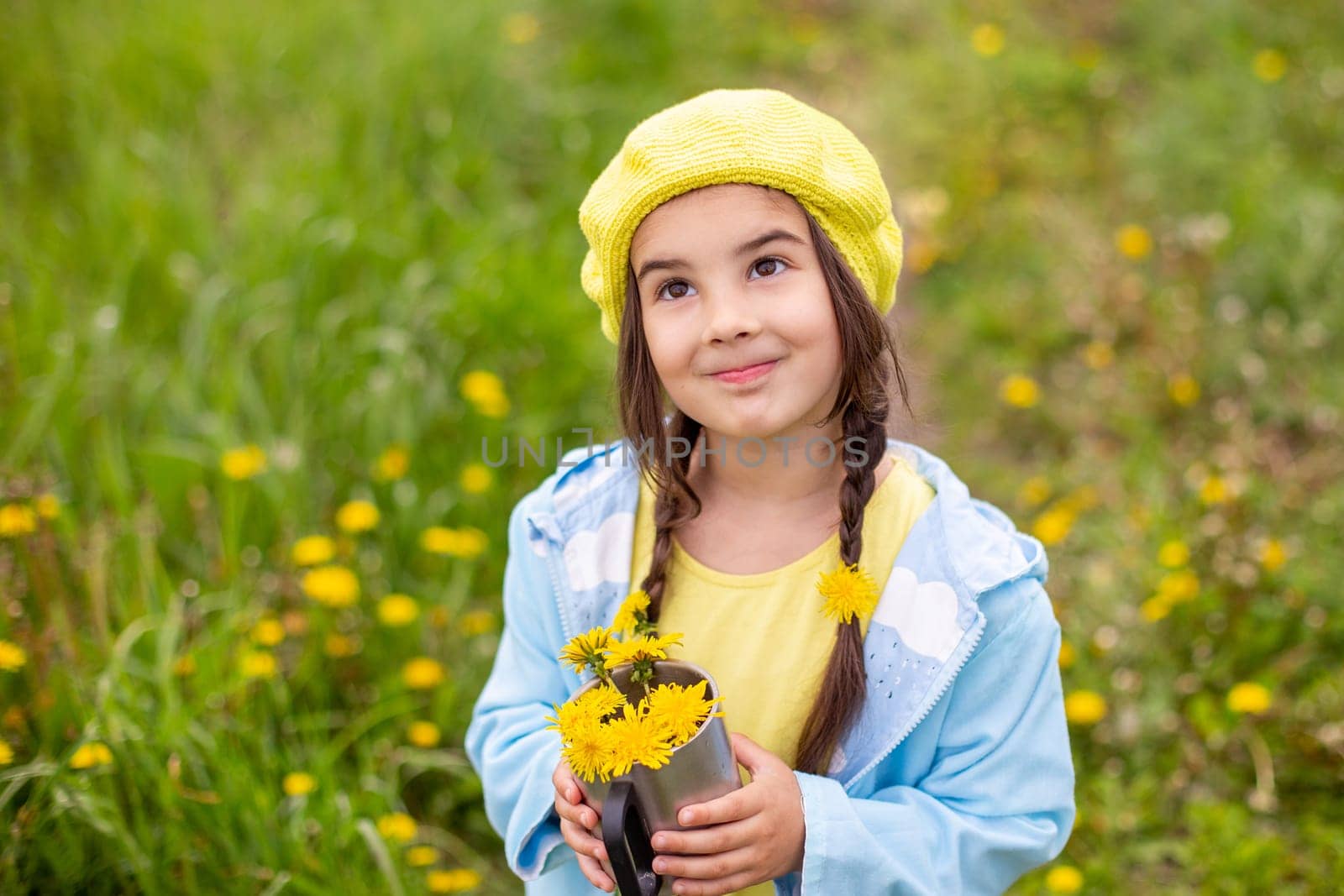 Portrait of a beautiful little girl with two pigtails, in a yellow beret, blue raincoat, stands in nature on a green lawn with dandelions, smiles, holds a bouquet of yellow dandelions in a metal mug