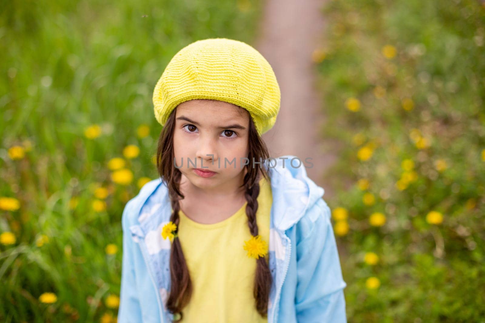 Portrait of a sad adorable little girl with two pigtails with braided dandelions, in a yellow beret, blue raincoat, stands in nature, a green lawn with dandelions, looks at the camera.