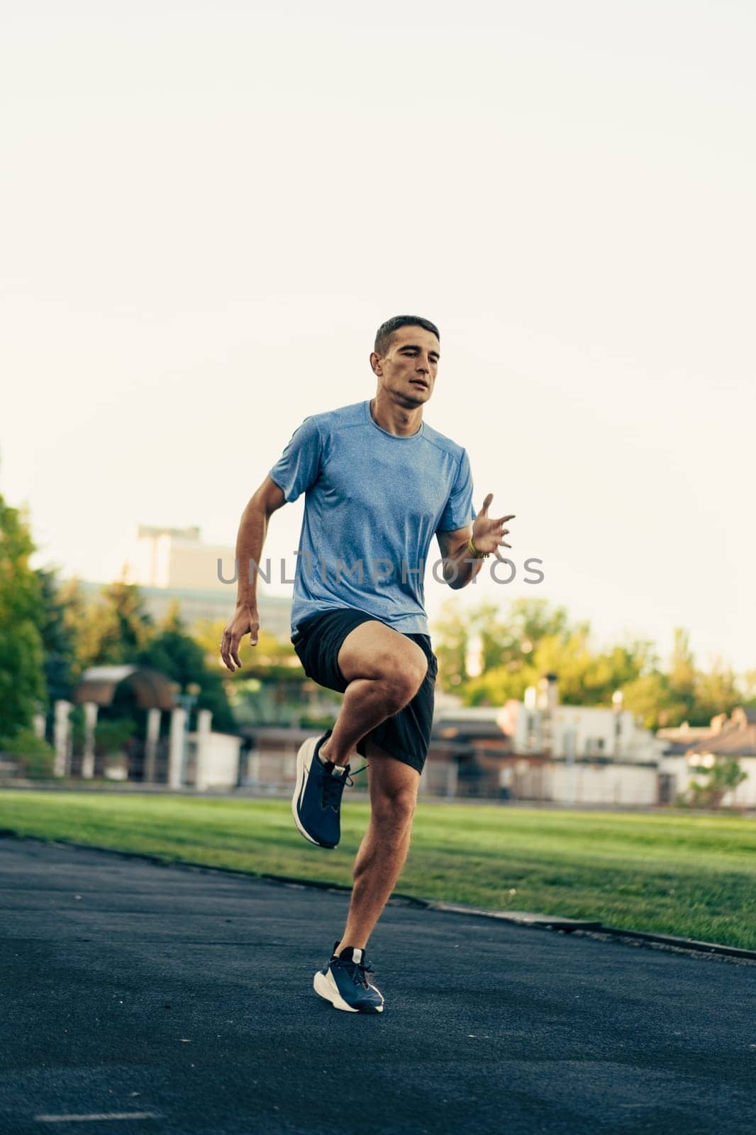 A man exercises high knees on a track, showcasing his speed and agility. The sun rises in the background, illuminating the training facility and surrounding greenery.