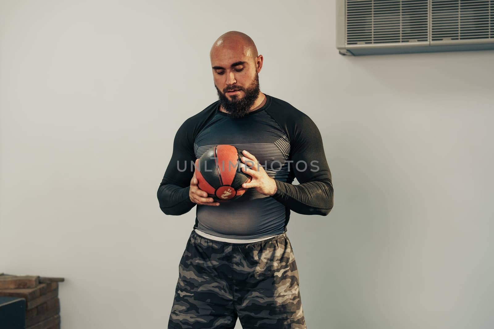 A man engages in strength training at a gym, holding a medicine ball with focused concentration. He is dressed in a fitted athletic shirt and camouflage shorts.