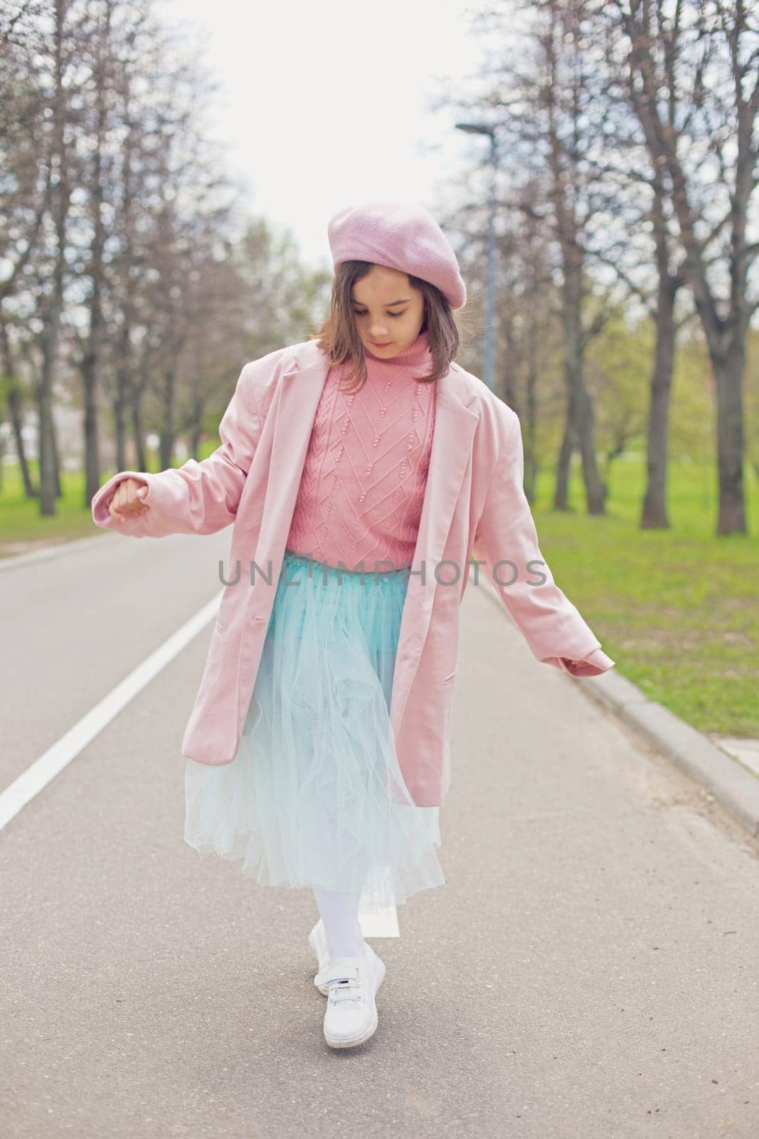 a little charming girl in a pink jumper, jacket, beret and light blue skirt walks along an asphalt road in the spring park. Vertical