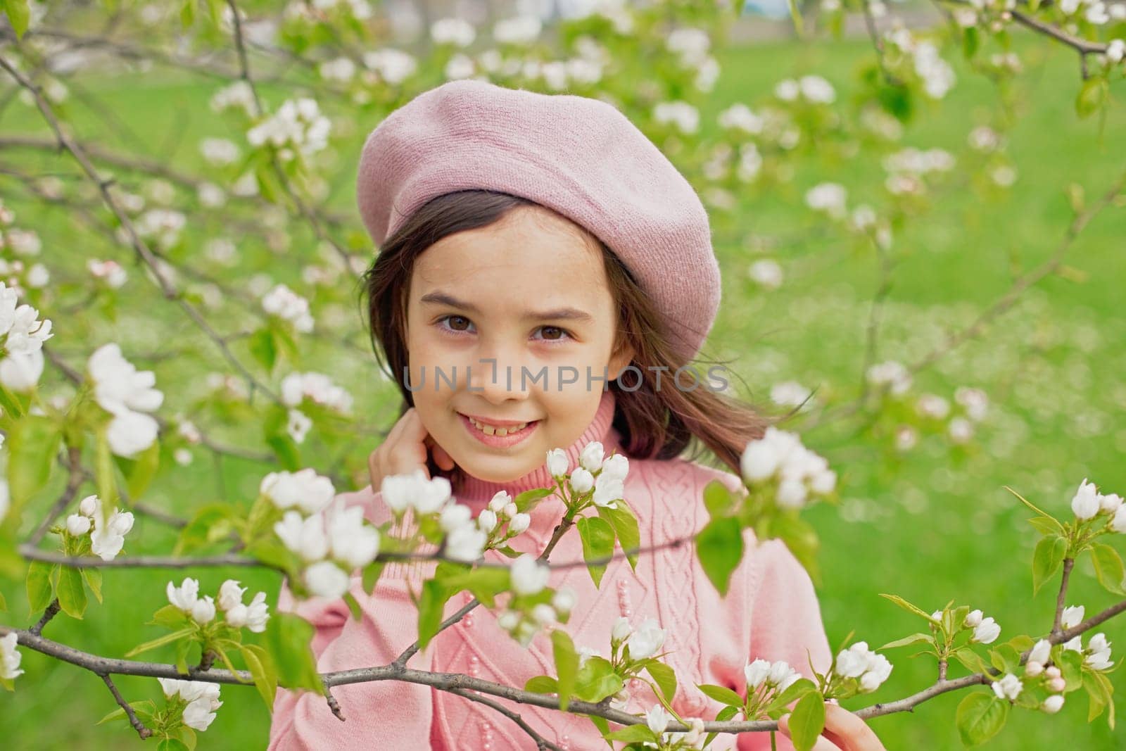 Portrait of a beautiful little girl in a pink jumper and beret stands next to the branches of a blossoming white apple tree in the park. Close-up