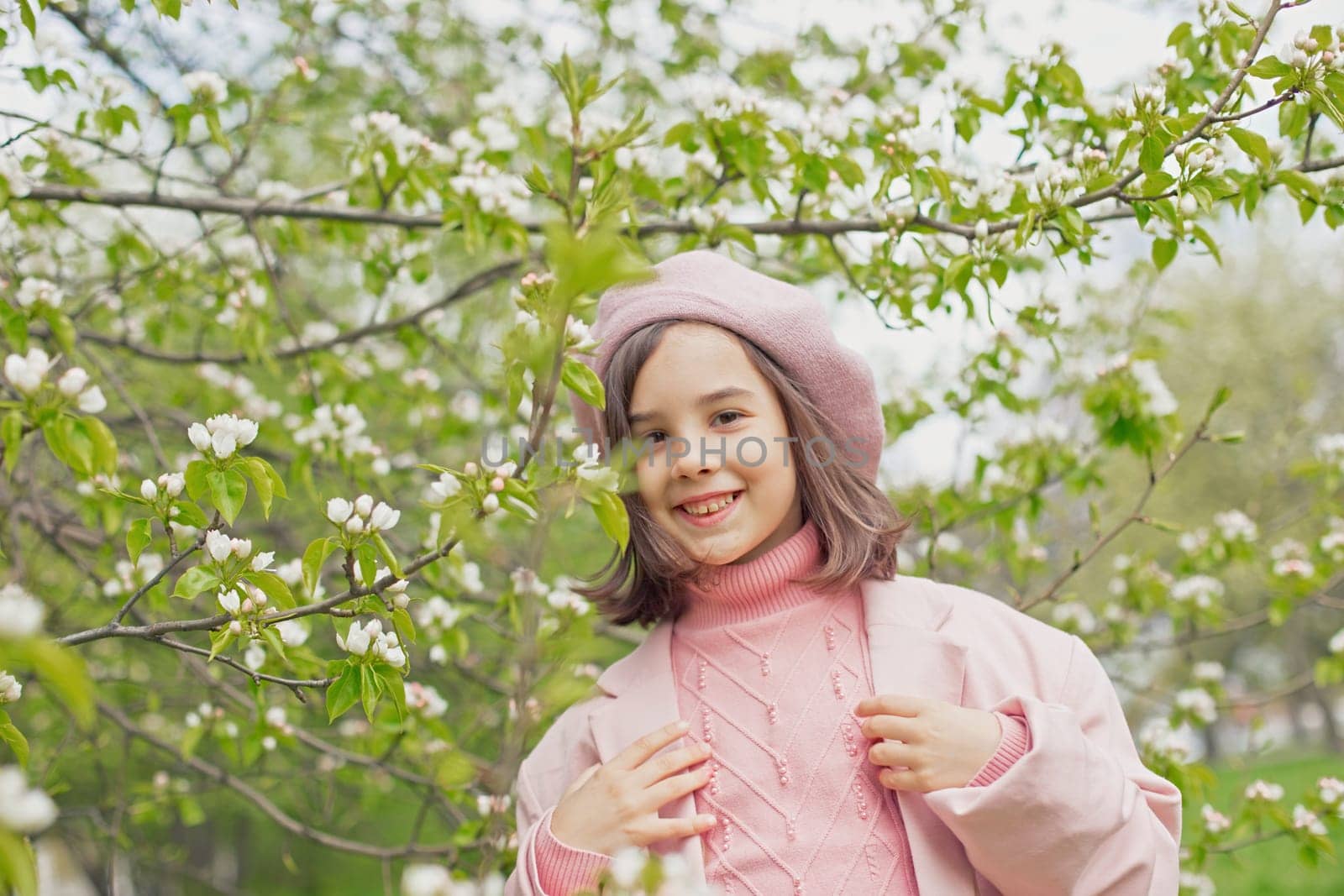 Portrait of a happy beautiful little girl in a pink jumper and beret stands next to the branches of a blossoming white apple tree in the park. Close-up
