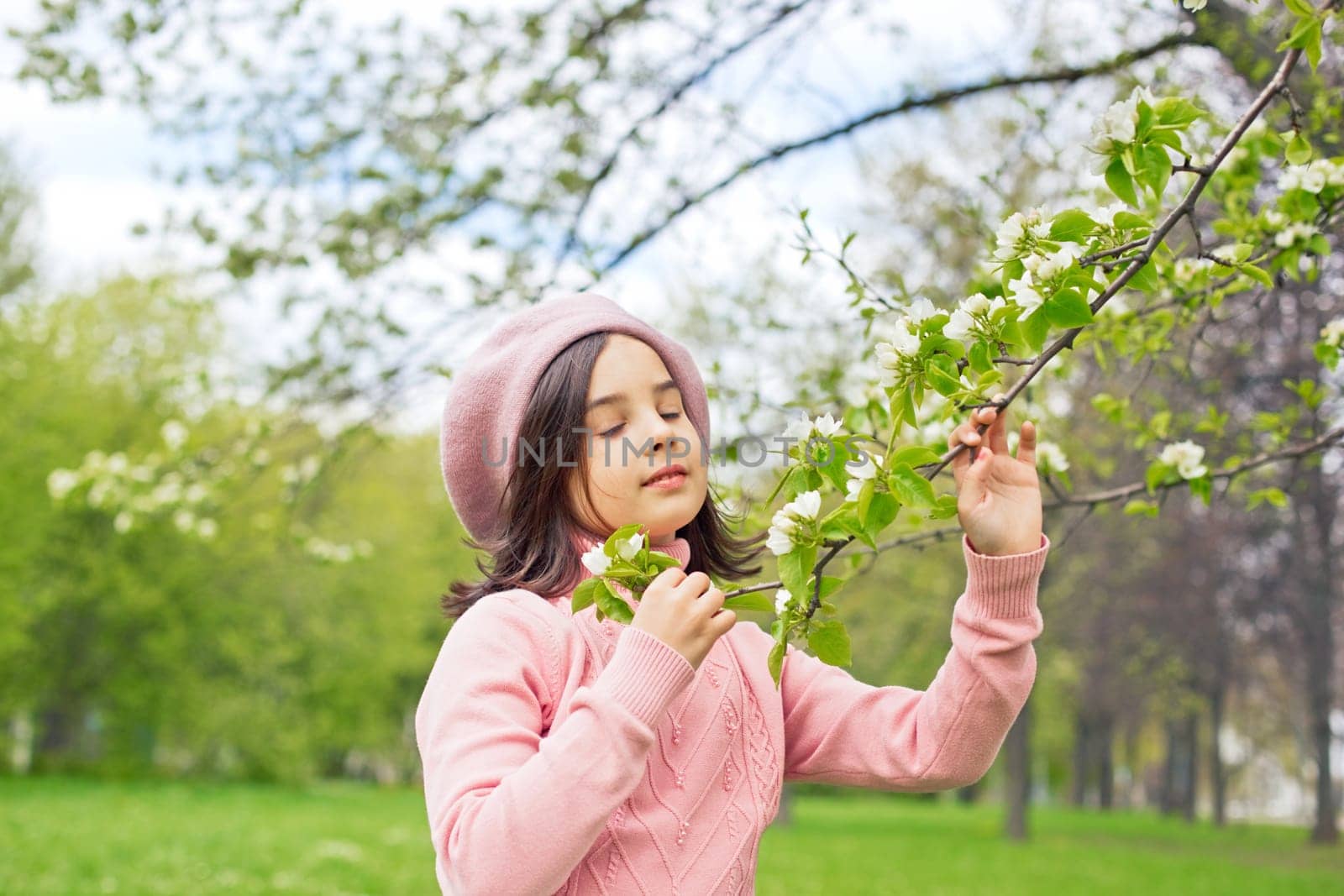 A beautiful little girl in a pink jumper holds a branch of a blossoming white apple tree in the park during the day, closing her eyes. Close-up