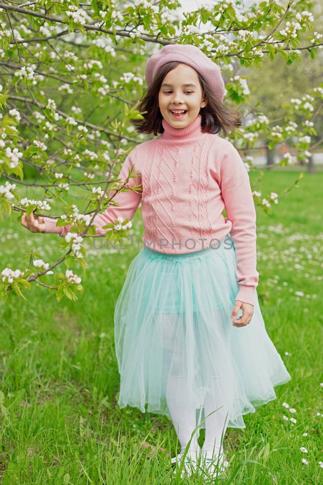 a happy little girl in a pink jumper, beret and blue skirt, stands next to a blooming white apple tree in the park. Close-up. Vertical