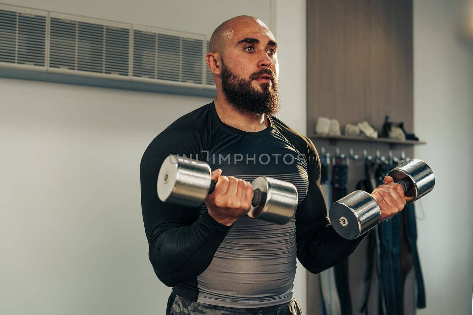 A man with a beard engages in strength training using two dumbbells in a well-lit gym. He focuses on his workout routine, indicating dedication to fitness and health.