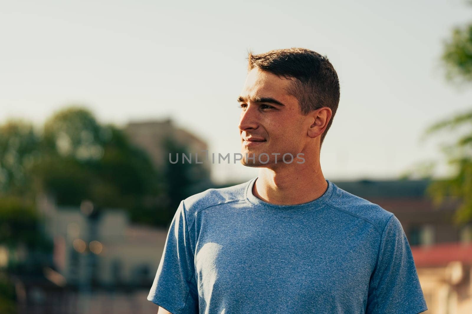 A young man stands outdoors in a city, wearing a blue athletic shirt. He gazes forward with a smile, enjoying a warm and sunny day, surrounded by buildings and greenery.