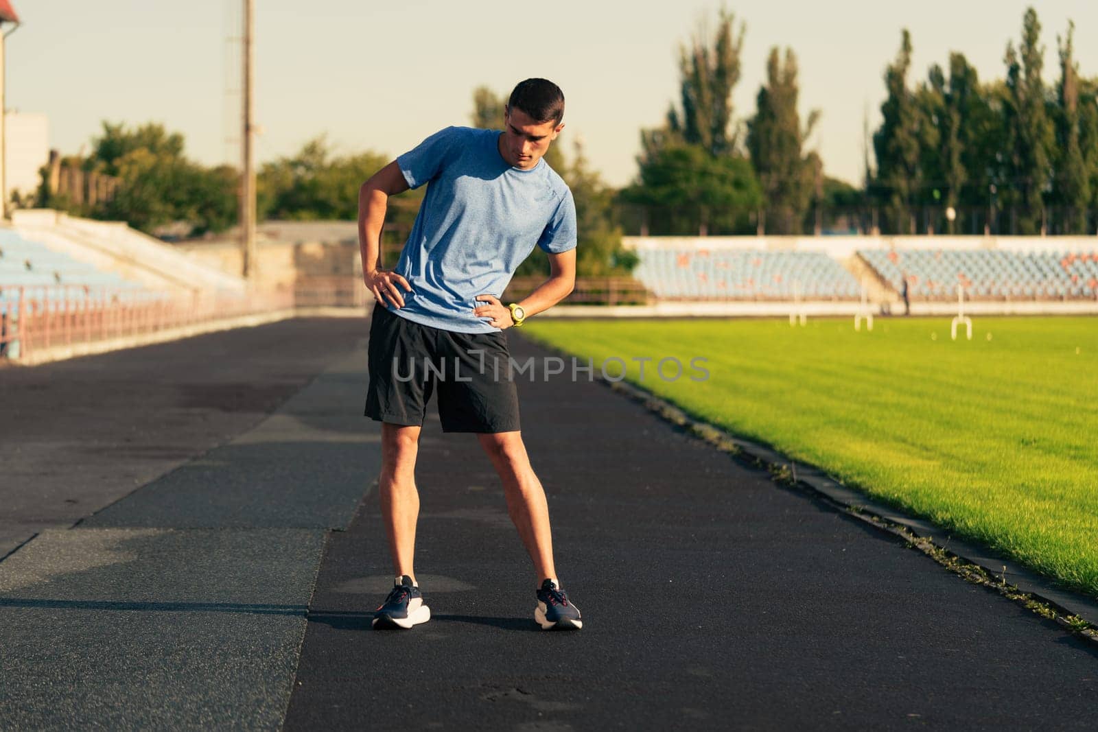 A young male athlete stretches his legs on a track field surrounded by green grass and empty bleachers, preparing for a running workout during the evening light.