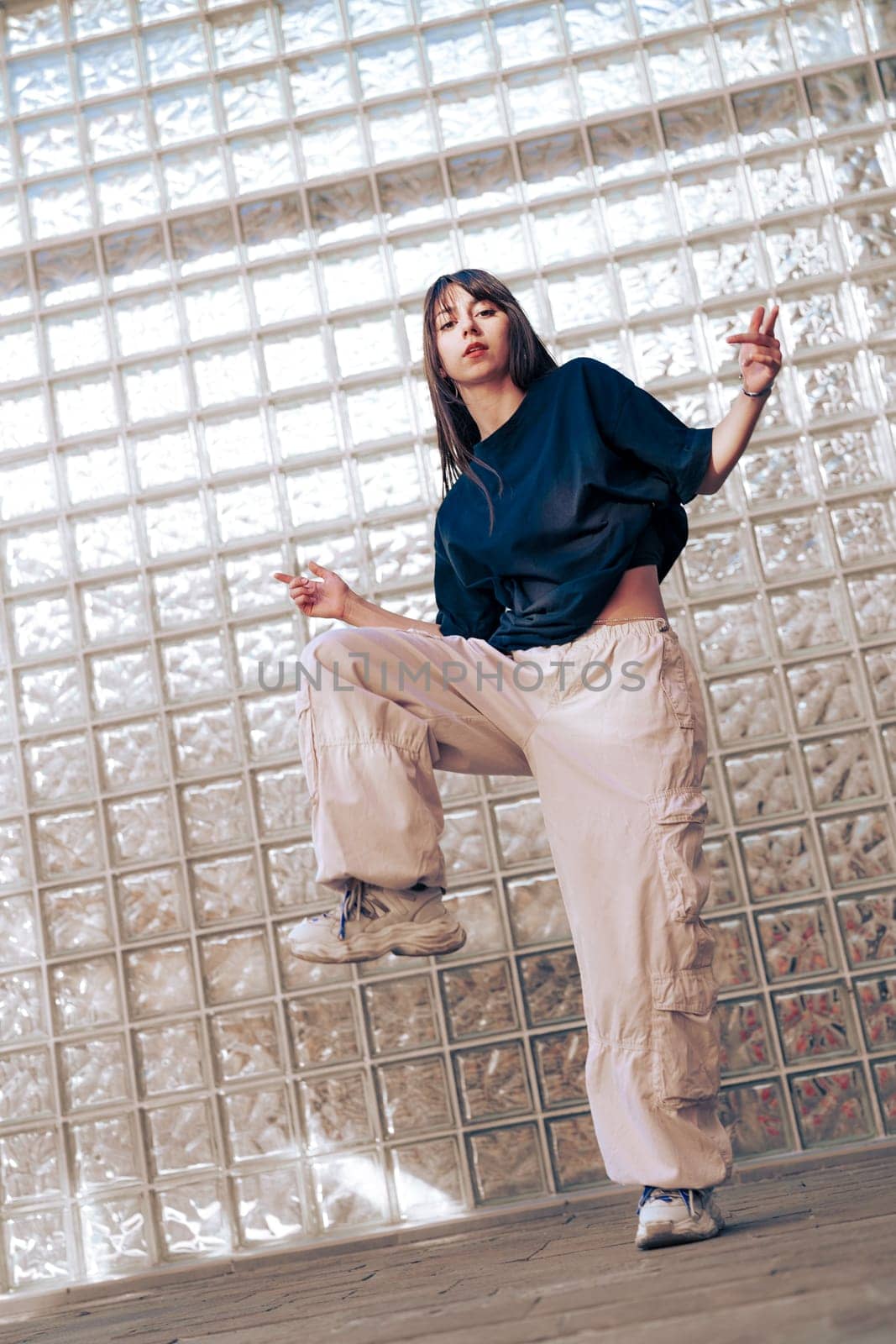 A dancer is performing a dynamic move indoors, wearing loose, stylish clothing. The background features a glass block wall that adds to the modern aesthetic of the scene.
