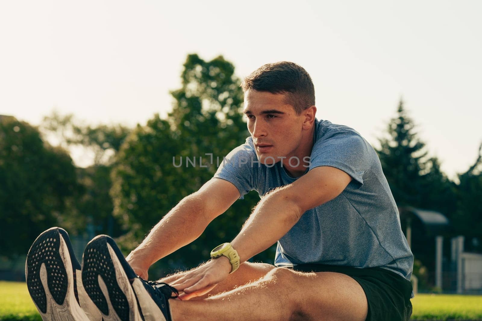 A young man stretches on green grass in a park during the day, focusing on fitness and preparation for his workout routine under clear skies.