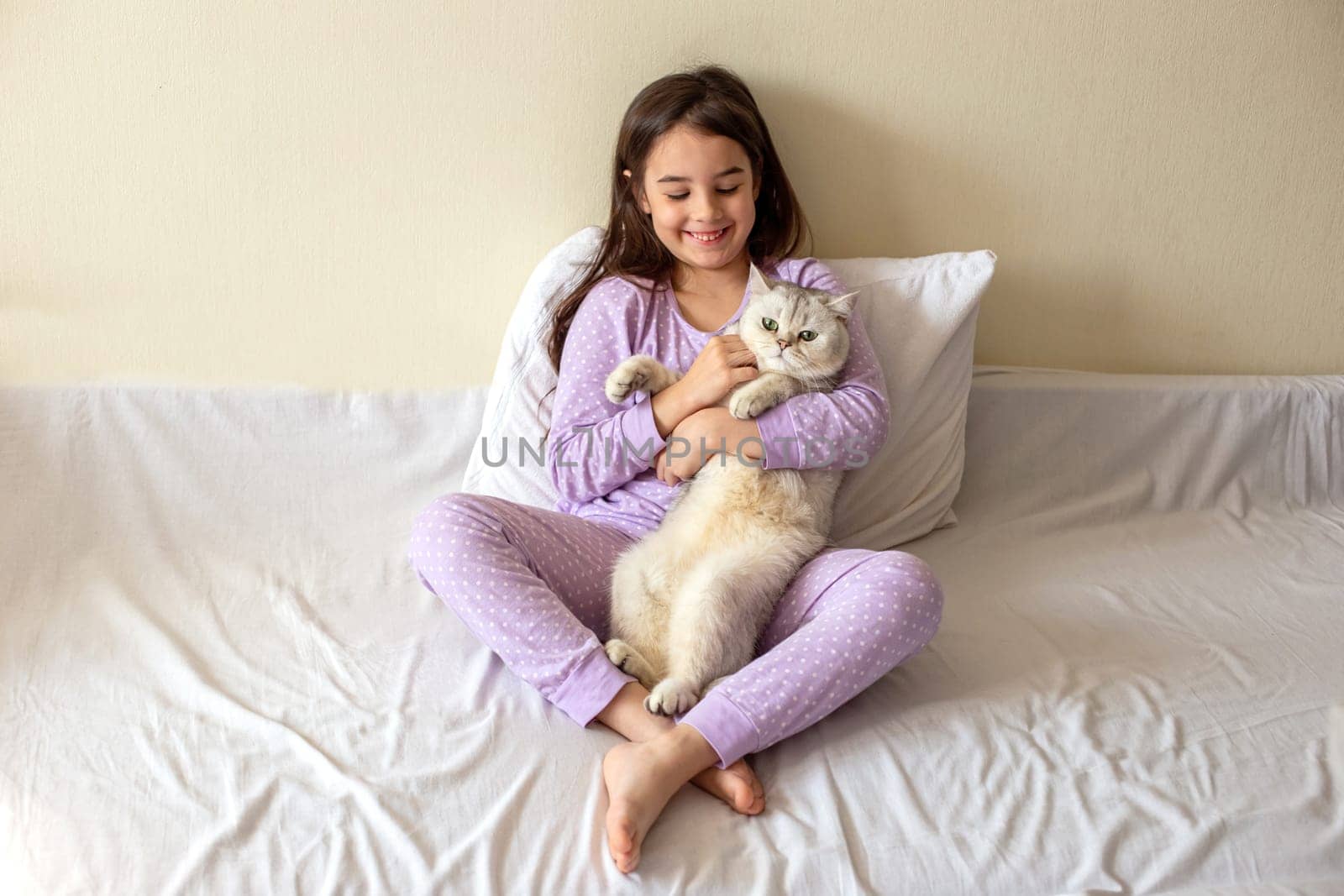 A happy little brunette girl in purple pajamas, with her dark hair loose, sits hugging a white charming cat on a white bed, smiling