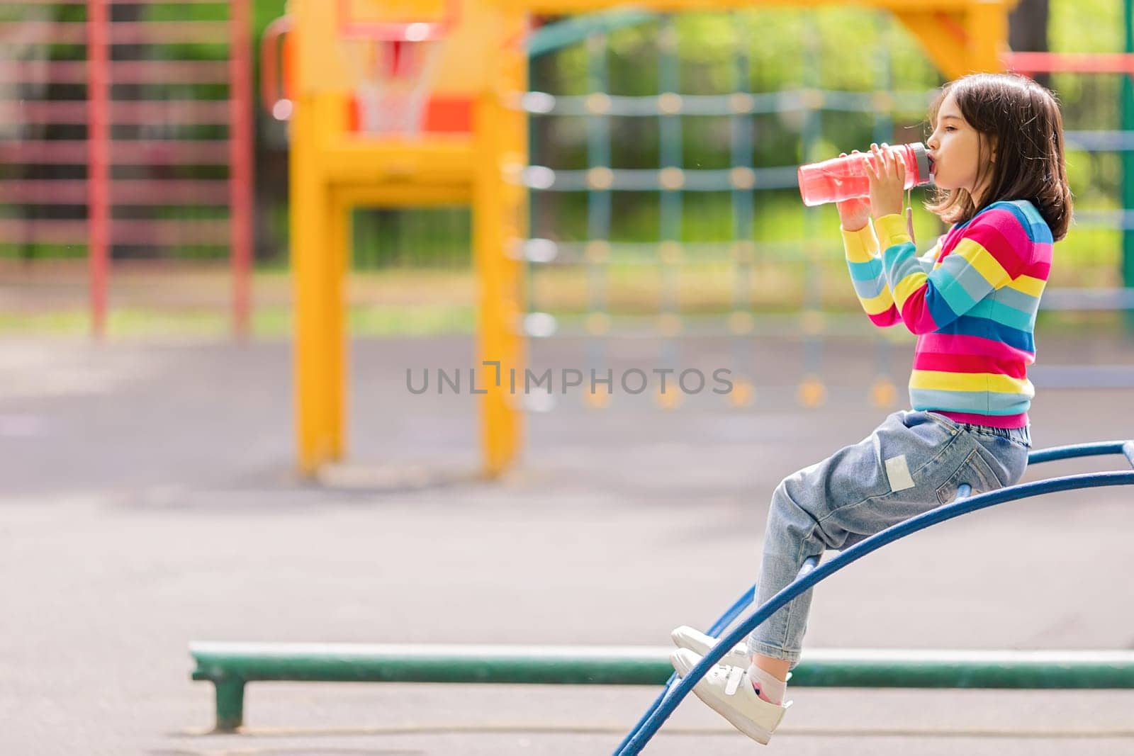 Little girl 5-6 years old, sits on the playground in summer, drinks water and a pink plastic bottle. Copy space