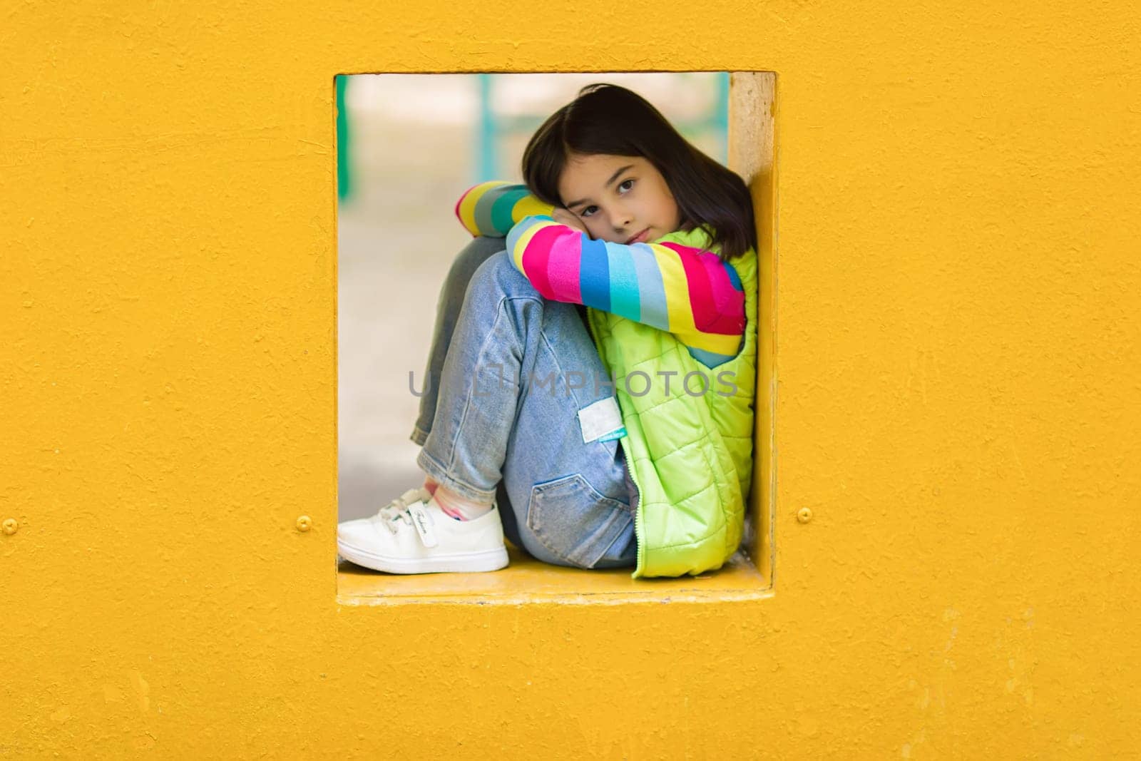 Sad and bored cute little girl is sitting among yellow colorful playground in summer. Close. copy space