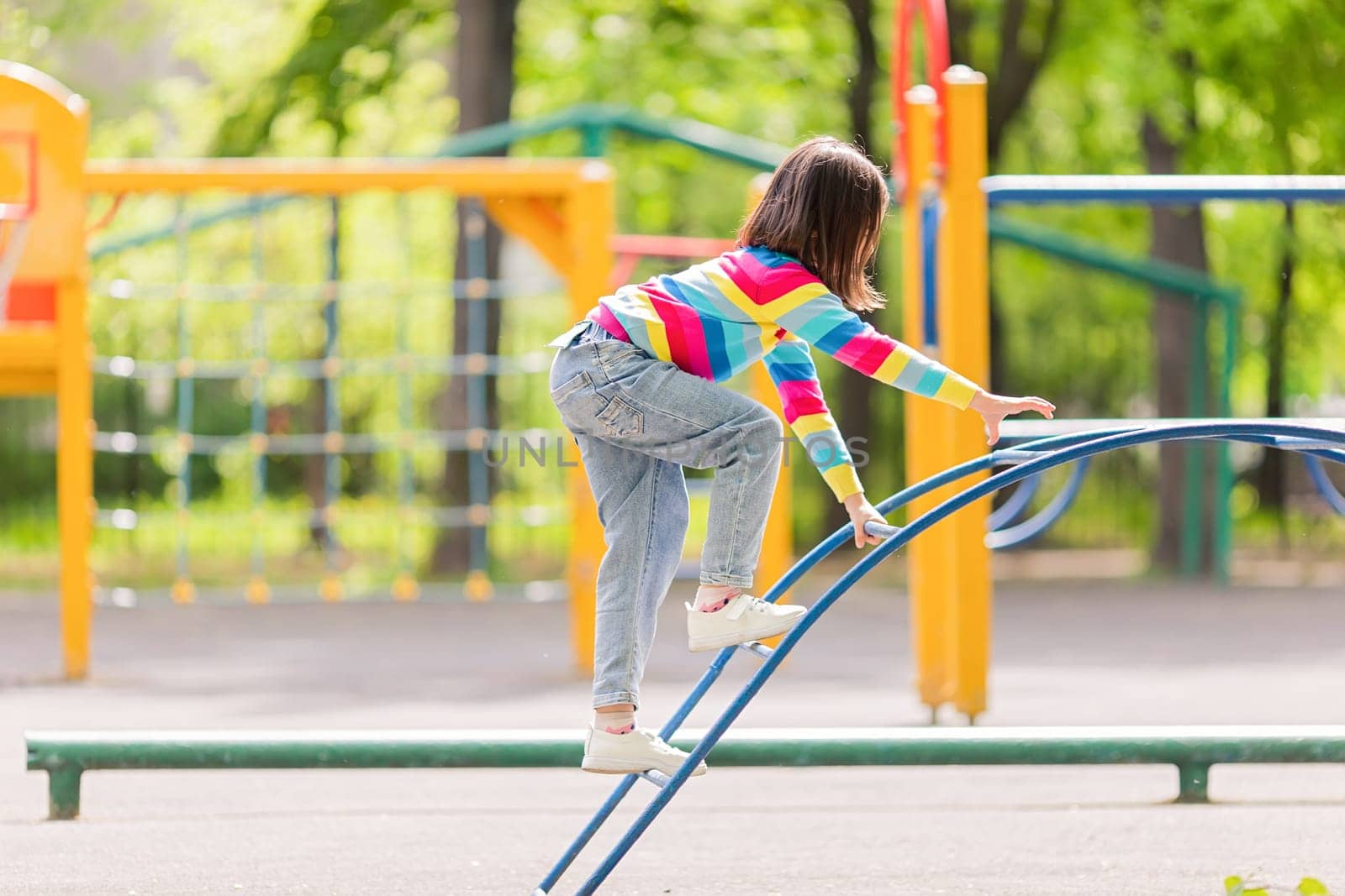 a girl in a bright striped jumper and jeans, climbs the childrens stairs on the playground in the park, in summer