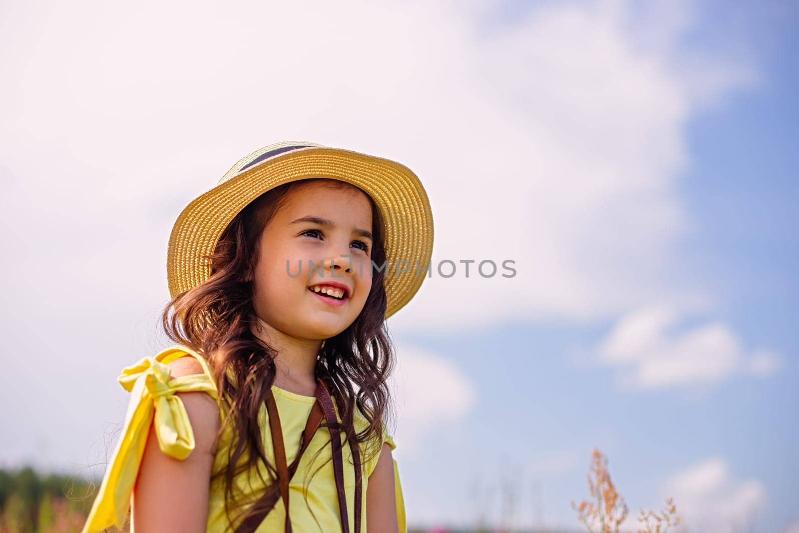 A happy cute little girl of 4-5 years old, in a straw hat and a yellow T-shirt, stands in nature in the summer, against the background of a blue sky, looks away