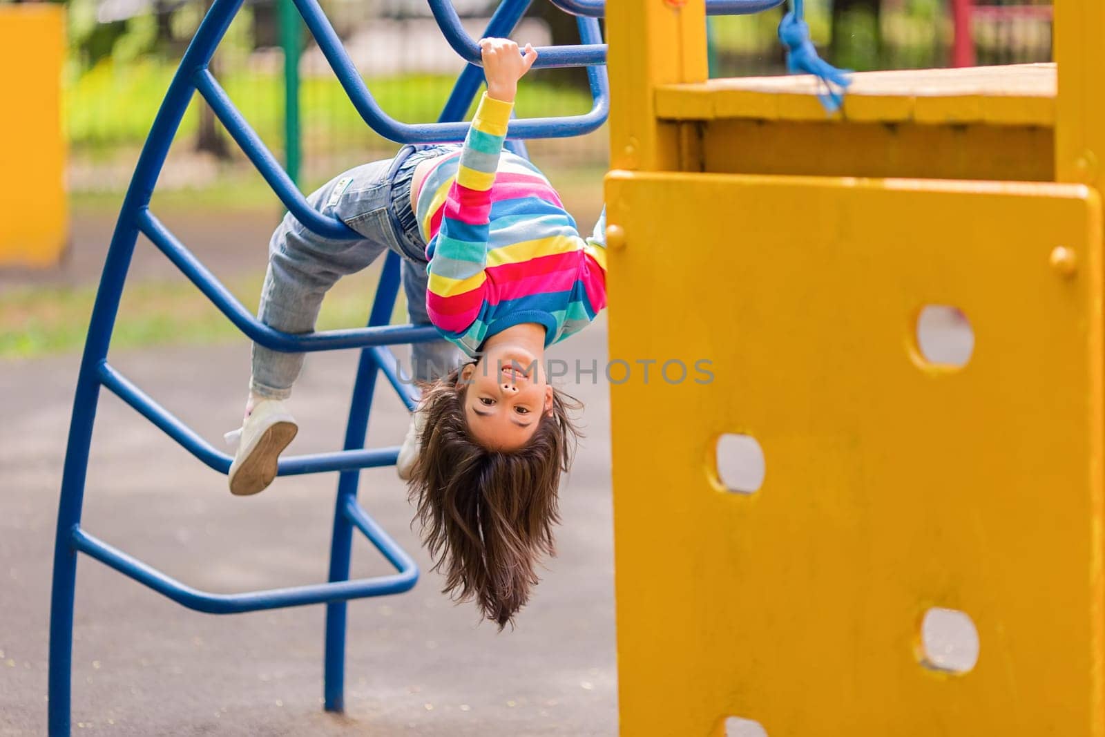 Funny little smiling girl 5-6 years old, hanging upside down on the children's stairs at the playground, in the park, in summer.Copy space