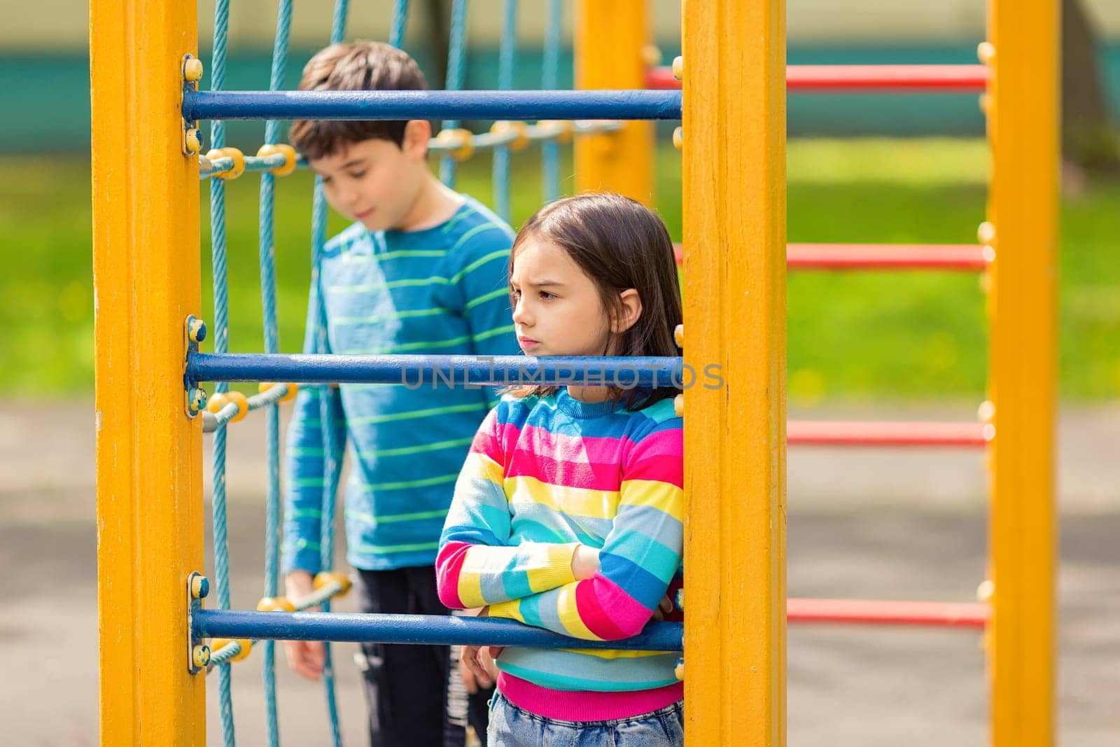 Sad and bored girl and boy stand among the yellow colorful playground, in the park, in summer, look away.