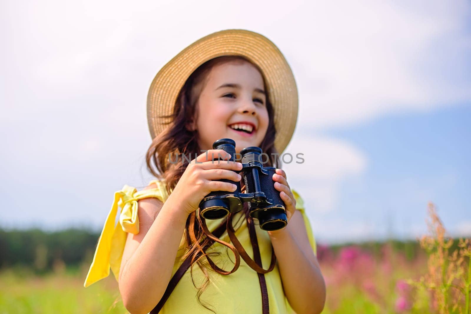 Happy cute little girl 4-5 years old, in a straw hat and yellow T-shirt, stands in nature in summer, against the background of the blue sky, holds binoculars