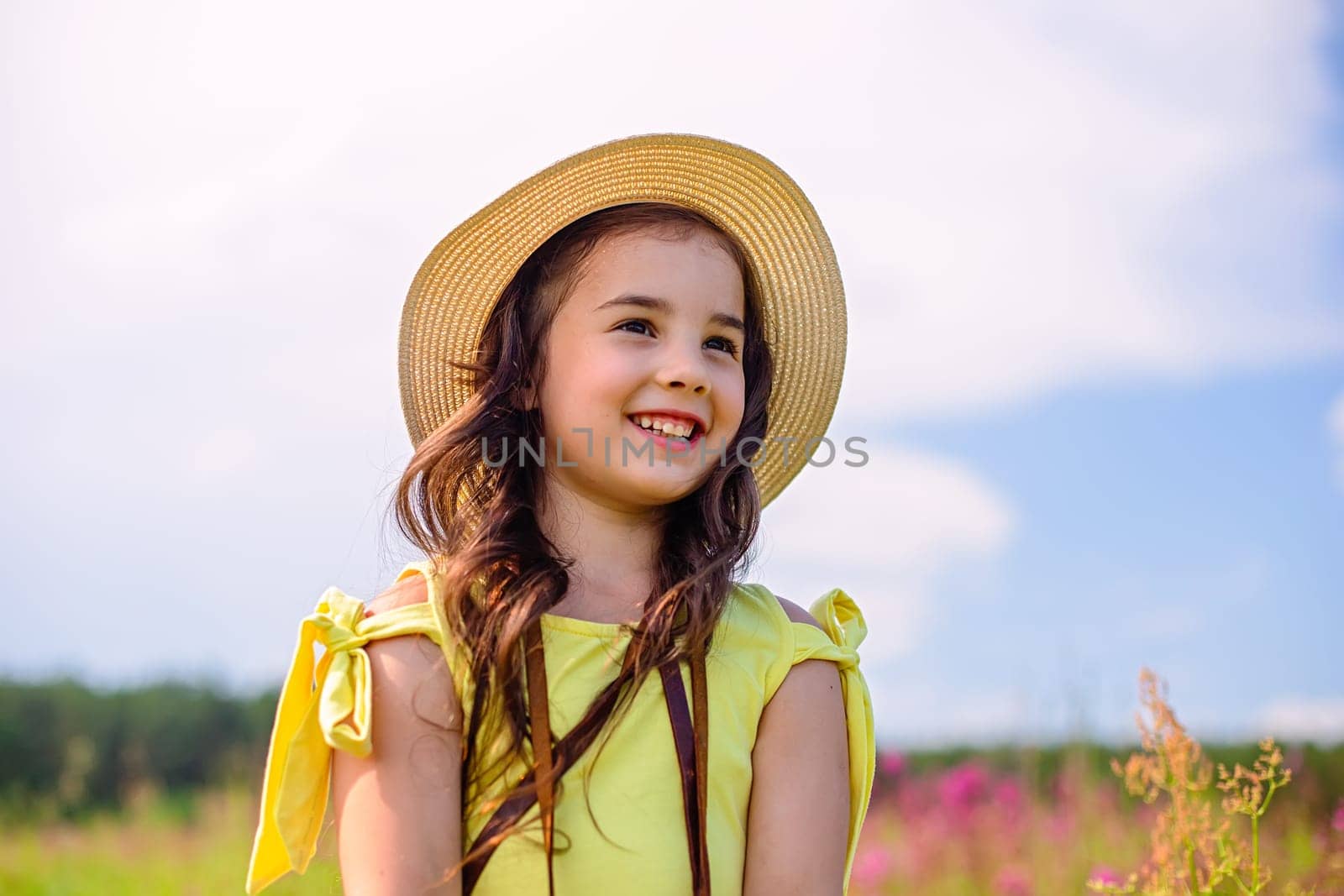 A smiling cute little girl in a straw hat and a yellow T-shirt, stands in nature in the summer, against the background of a blue sky, looks away