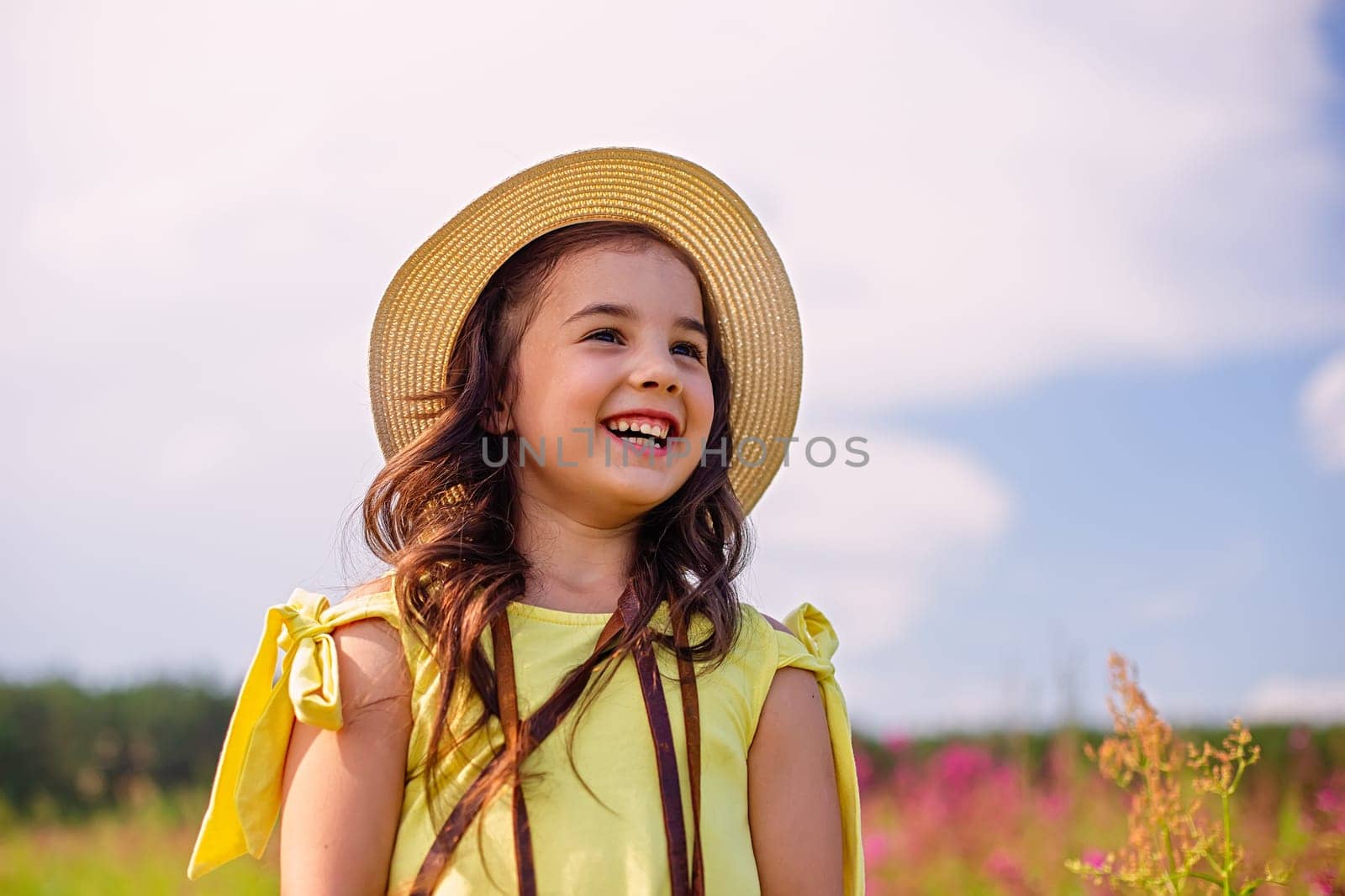 A happy brunette cute little girl in a straw hat and a yellow T-shirt, stands in nature in the summer, against the background of a blue sky, looks away
