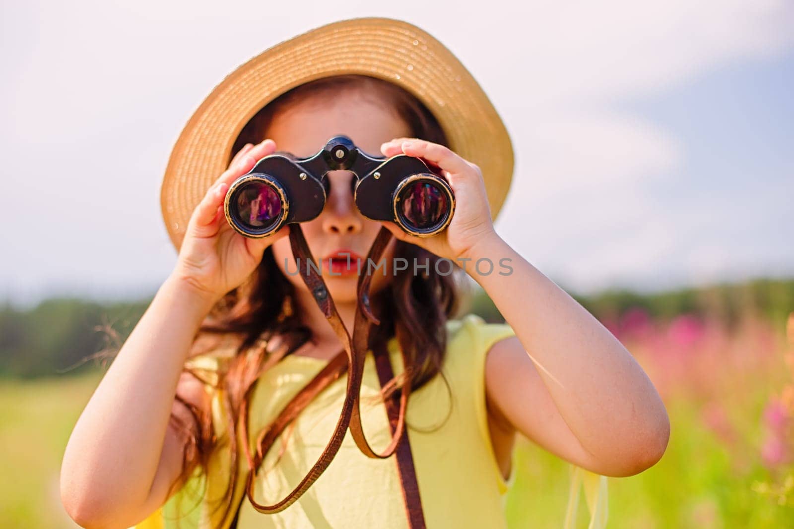 Surprised cute girl 4-5 years old in a straw hat and a yellow t-shirt in the summer stands in a field with tall grass against the blue sky, looks through binoculars. Close up