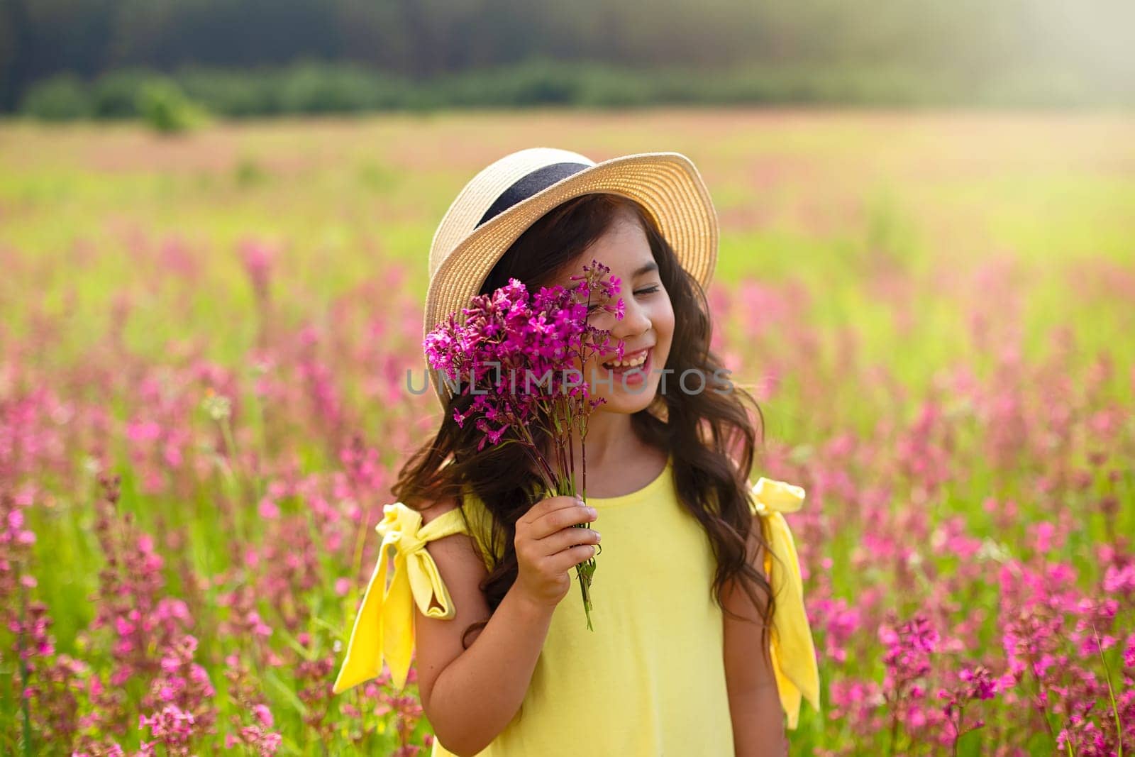 A beautiful playful little girl in a straw hat, with long dark hair, stands in a flowering field, in summer, holds a bouquet of burgundy viscaria flowers near her face. copy space. Close up