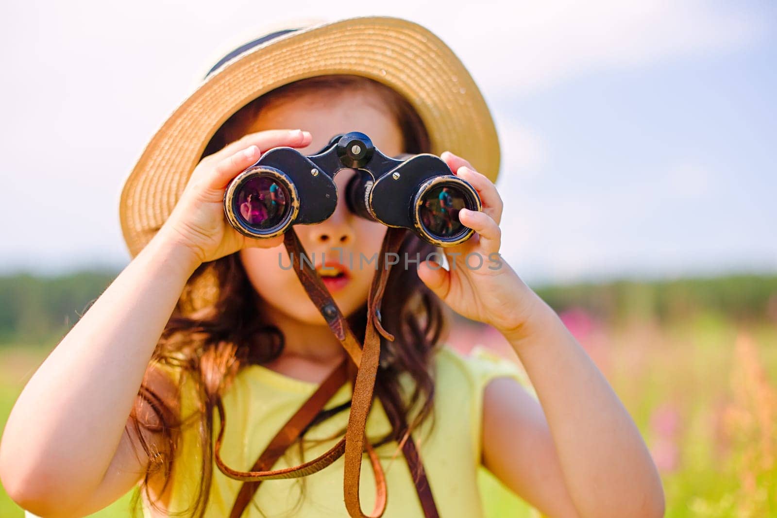 cute girl 4-5 years old, in a straw hat and a yellow T-shirt, in the summer, stands in nature, against the blue sky, looks through binoculars to the side