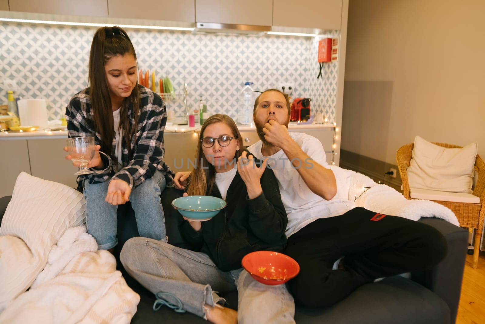 Three friends relax on a cozy couch, enjoying snacks and drinks in a warm, inviting home atmosphere