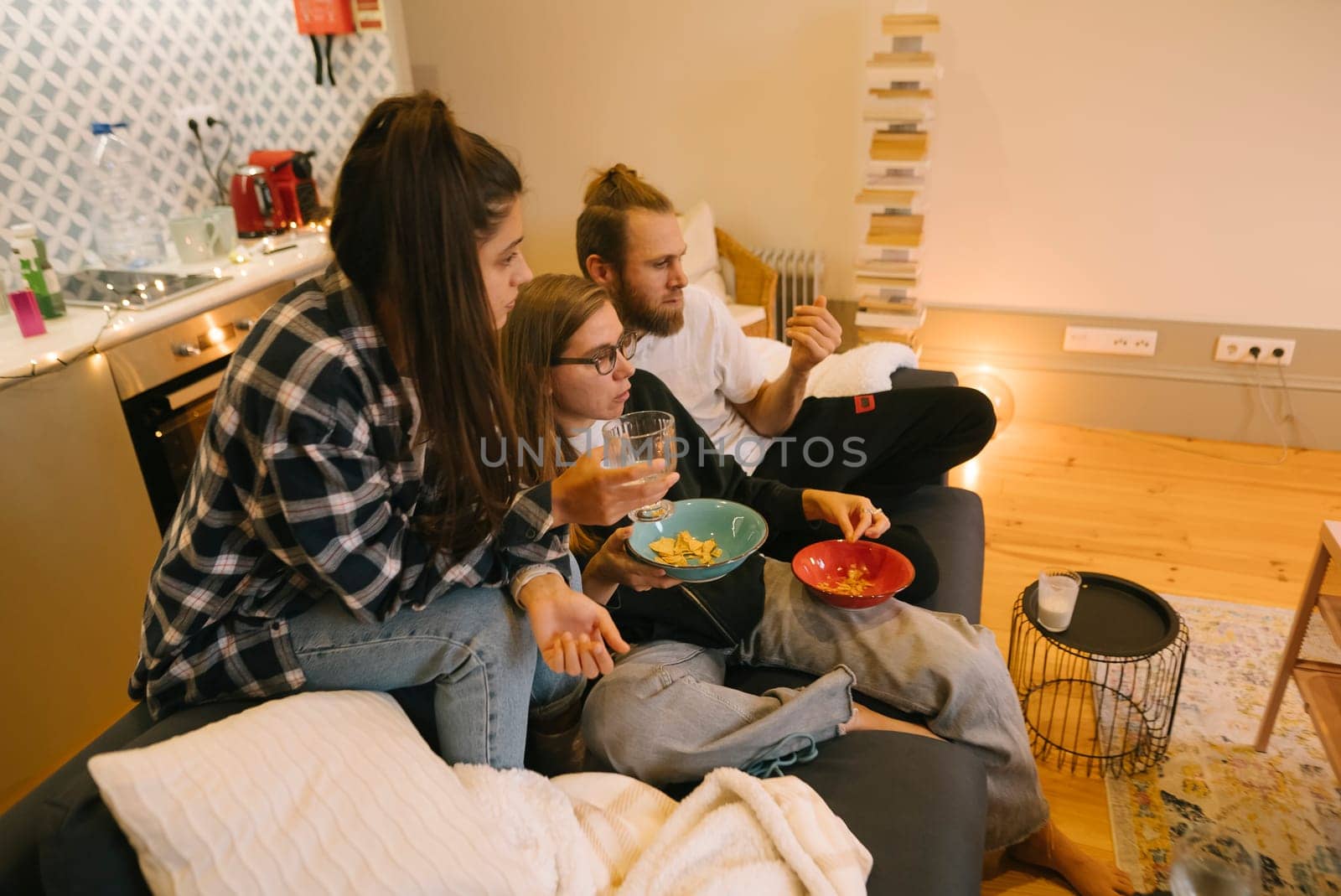 Three close friends are enjoying a cozy movie night at home together, filled with snacks, drinks, and laughter