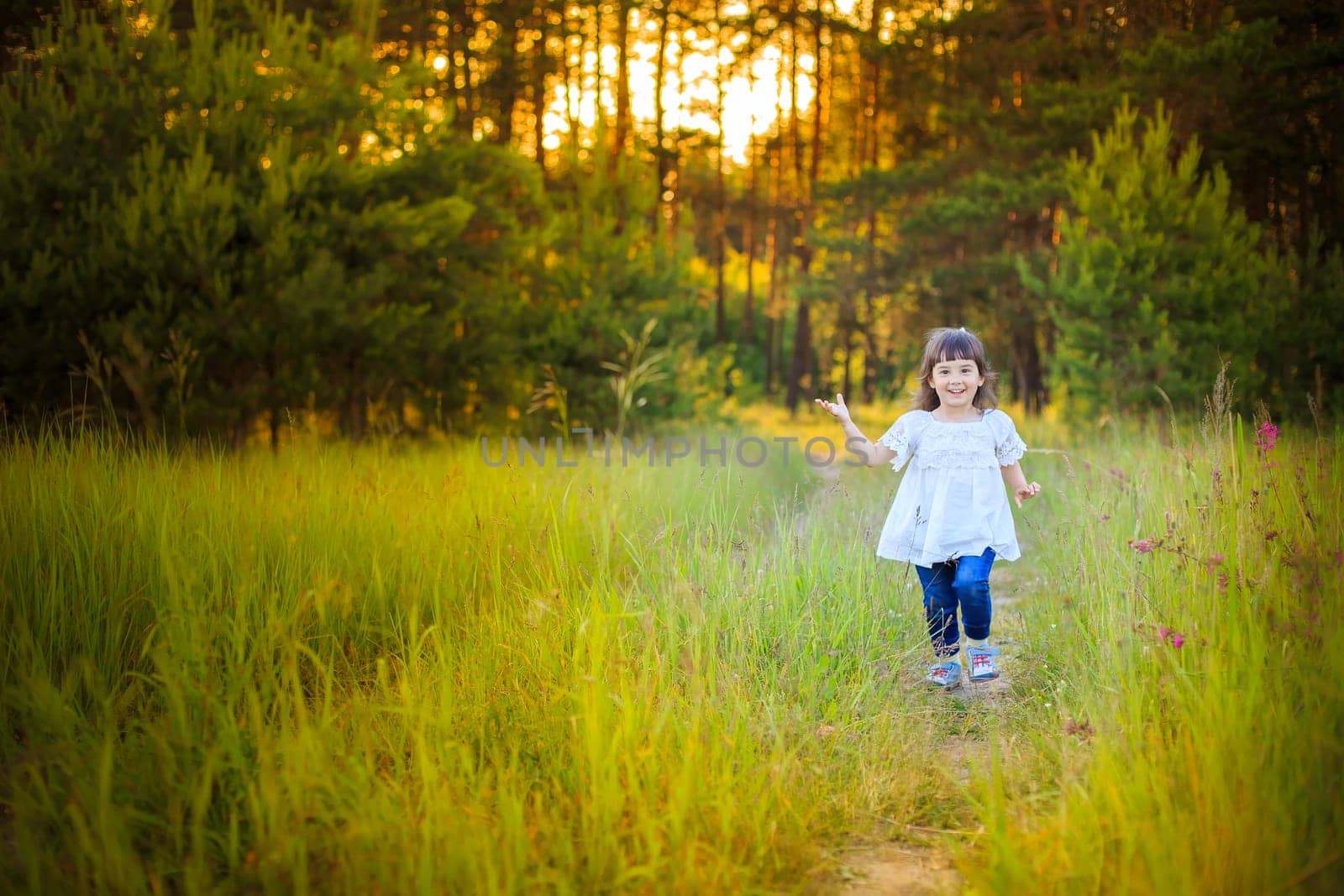 A charming happy little girl in a white dress walks along a path from a coniferous forest in summer,look at camera