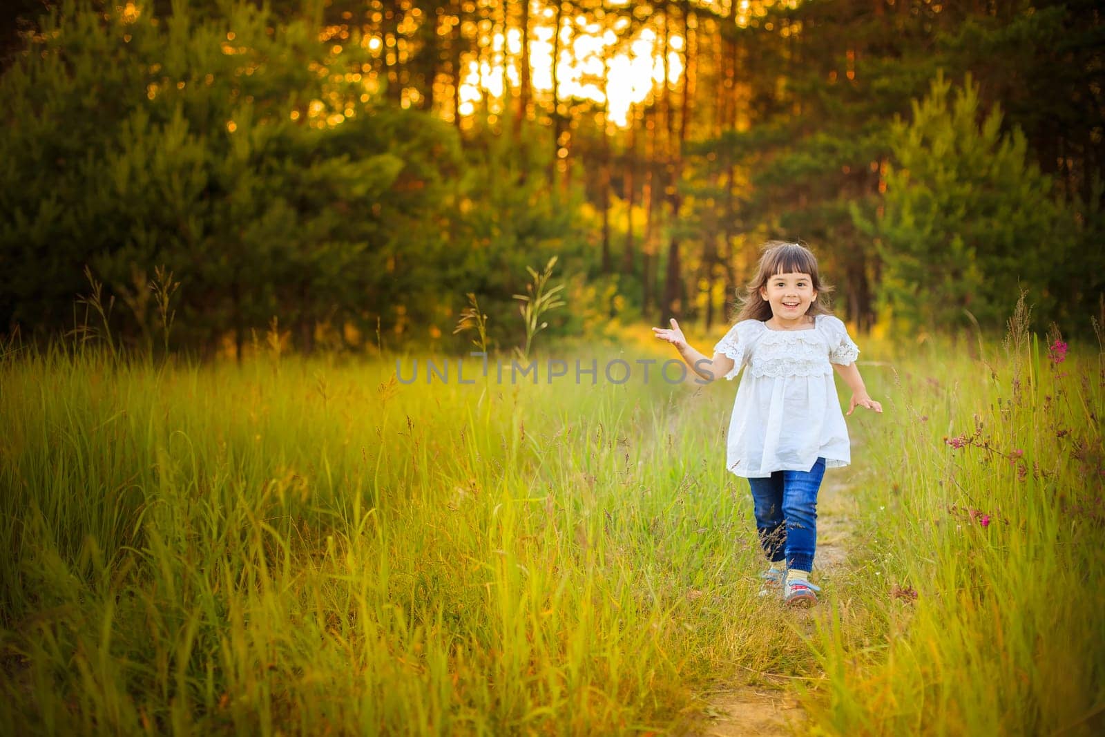 A happy little girl in a white dress runs along a path from a coniferous forest in summer, looks at the camera.