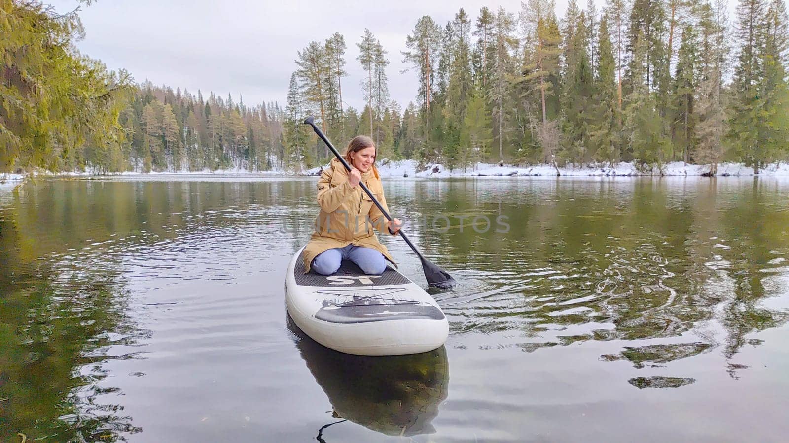 Kirov, Russia - March 14, 2024: A person paddles peacefully on a stand-up paddleboard in a serene lake, embraced by tall evergreen trees and a calm atmosphere.
