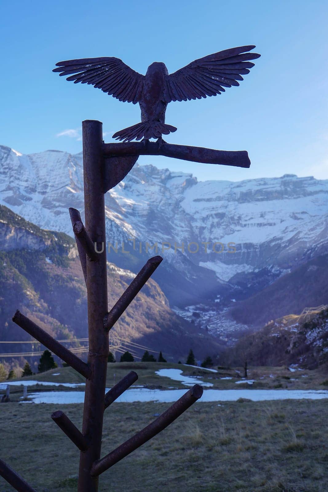 A detailed eagle sculpture perches on a branch, silhouetted against stunning mountain peaks in a tranquil valley. The scene captures the essence of nature's beauty.