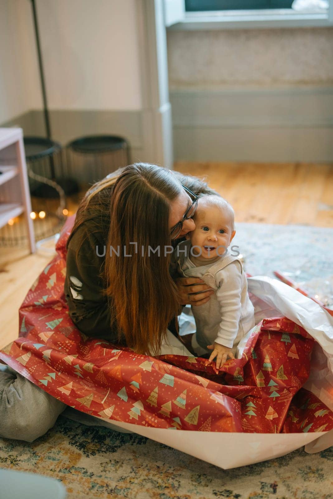 A mother joyfully interacts with her baby, both enveloped in a cheerful, festive sea of vibrant wrapping paper
