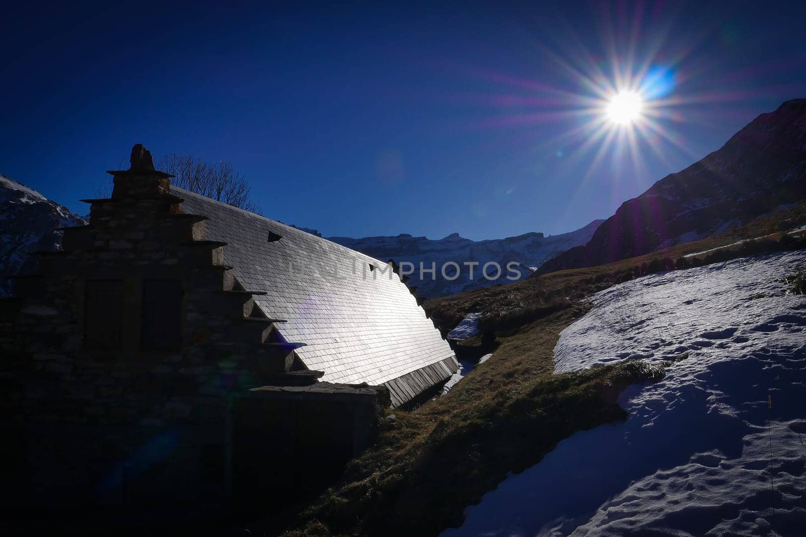 A rustic mountain hut rests on a lush hillside blanketed with snow, illuminated by a radiant sun in the clear blue sky. The scene captures the tranquil beauty of nature in the highlands.