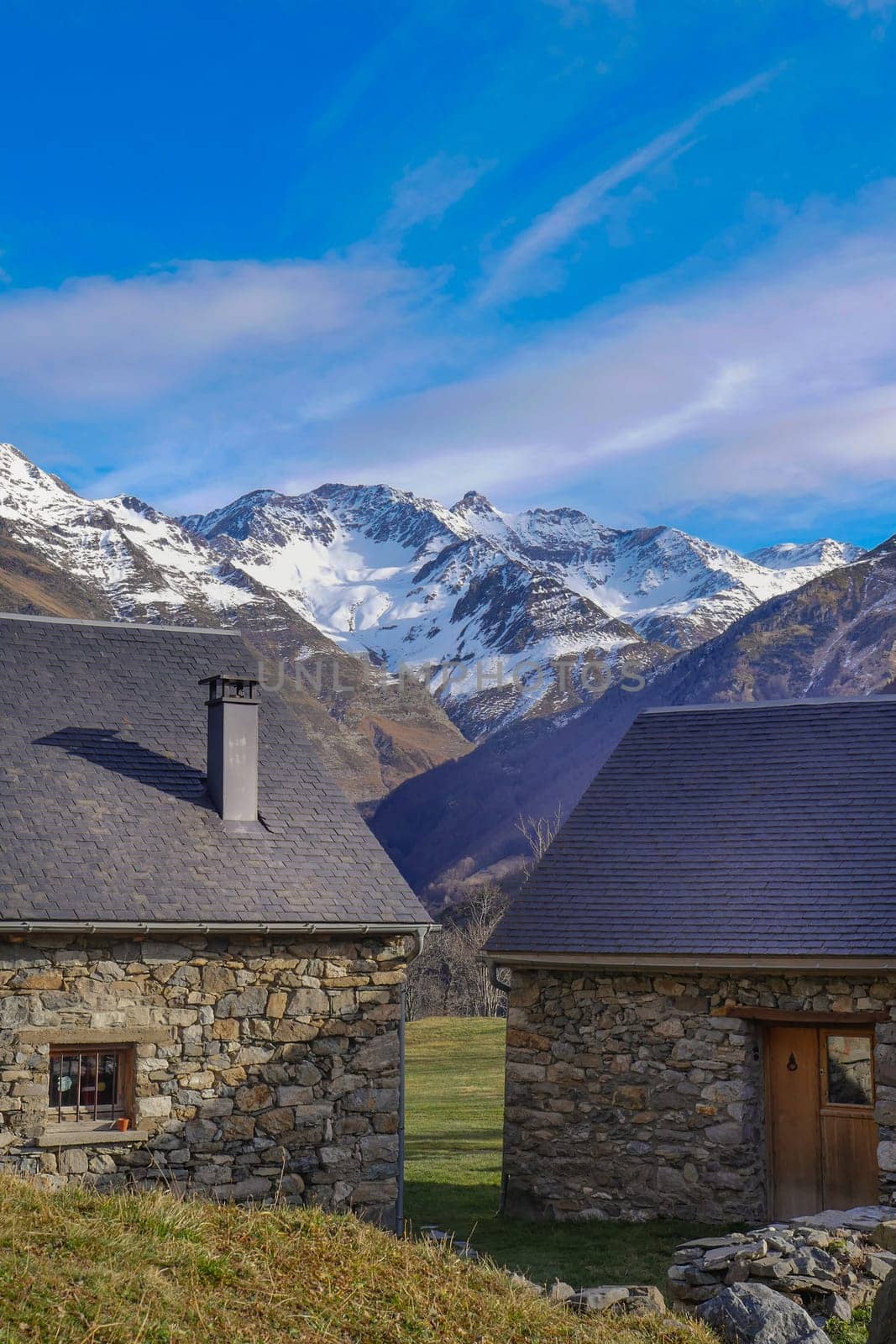 Two rustic stone cottages sit in a tranquil valley with snowy mountains rising majestically in the background under a bright blue sky, creating a serene escape for visitors.