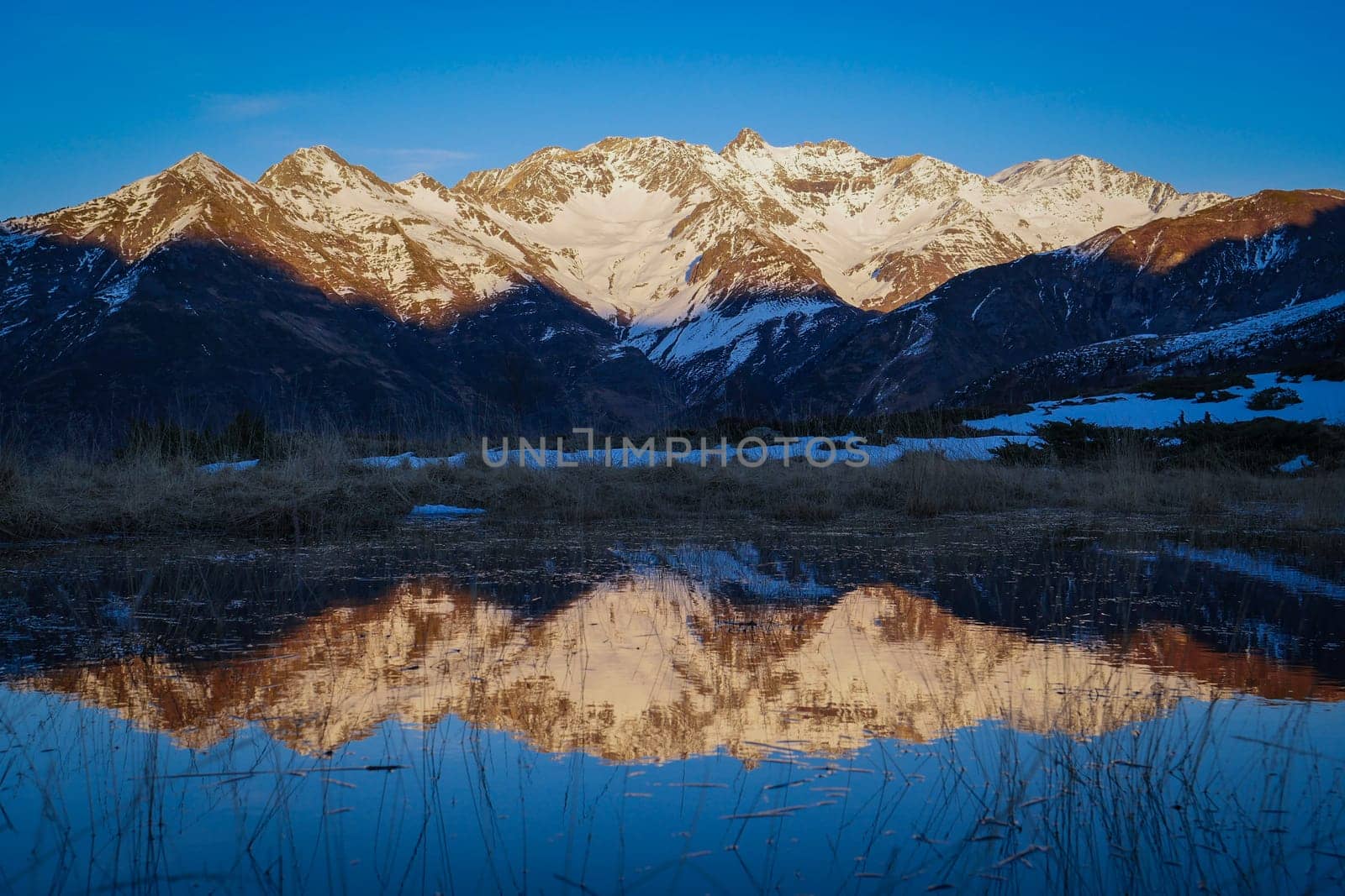 Snow-covered peaks rise majestically against a clear blue sky, with their reflection captured in still water at dawn in a peaceful alpine setting.