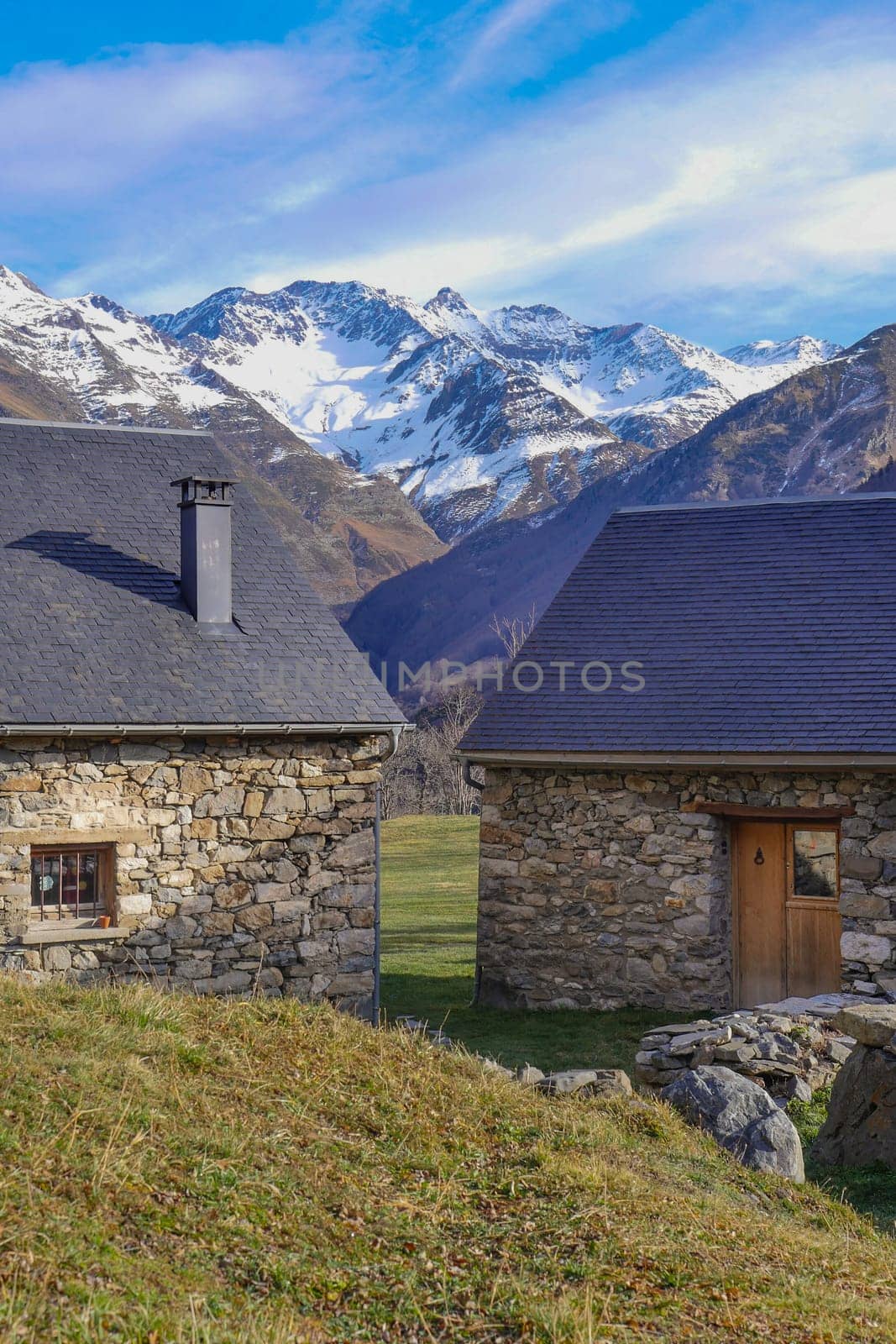 Two charming stone houses are nestled in a serene valley, surrounded by towering snow-capped mountains under a bright blue sky. The picturesque setting exudes tranquility.