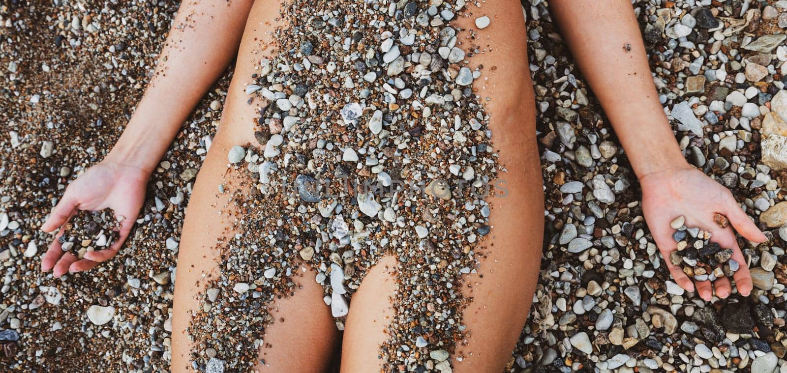 A nude woman reclines on a sandy beach, her body partially covered with small pebbles. She is relaxed, enjoying the warmth of the sun on a clear day.